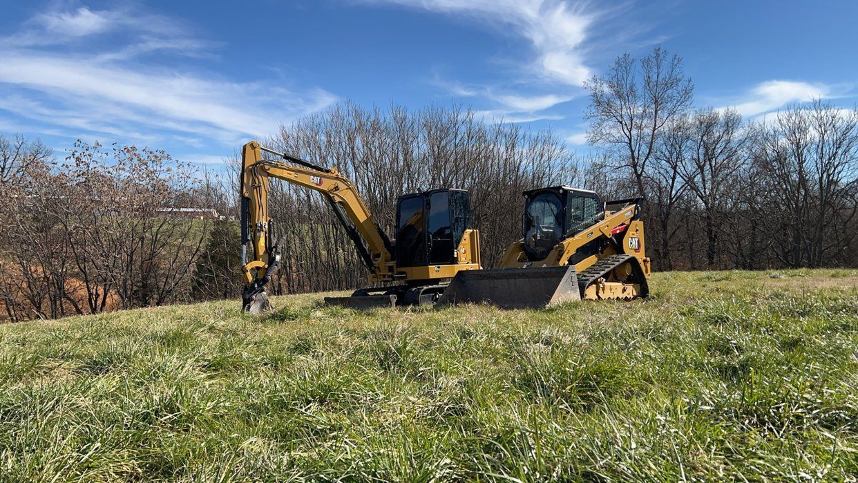 Yellow excavator and skid steer on a grassy hillside under a blue sky.
