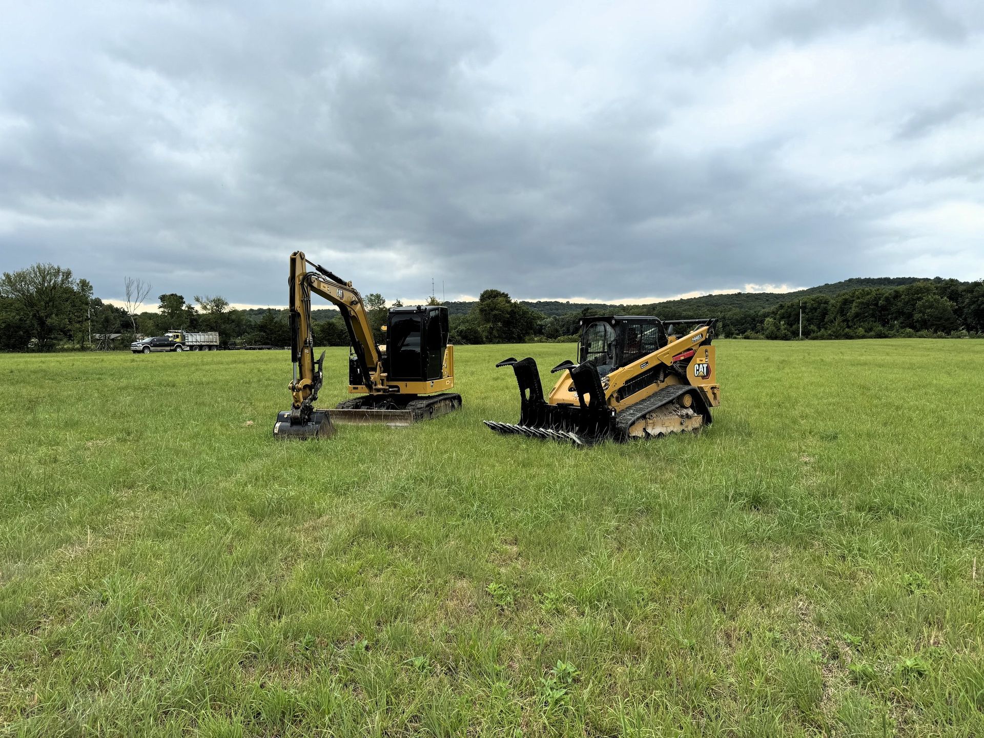Two yellow construction vehicles, an excavator and a skid steer, sit in a grassy field under a cloudy sky.