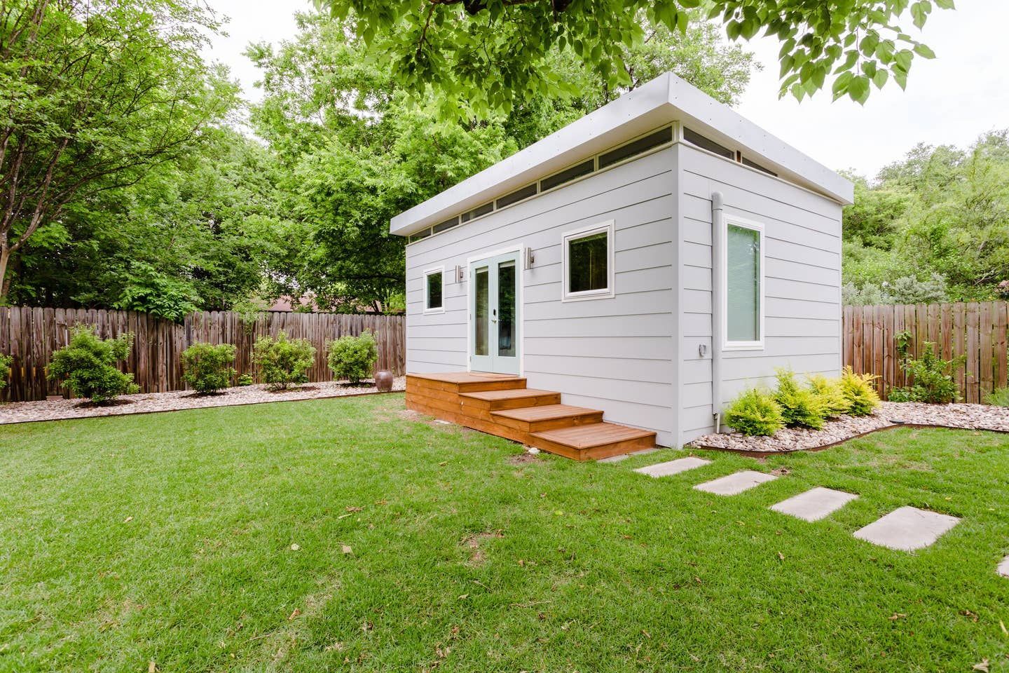 White modern shed with wood steps in a backyard with green grass, trees, and a wooden fence.
