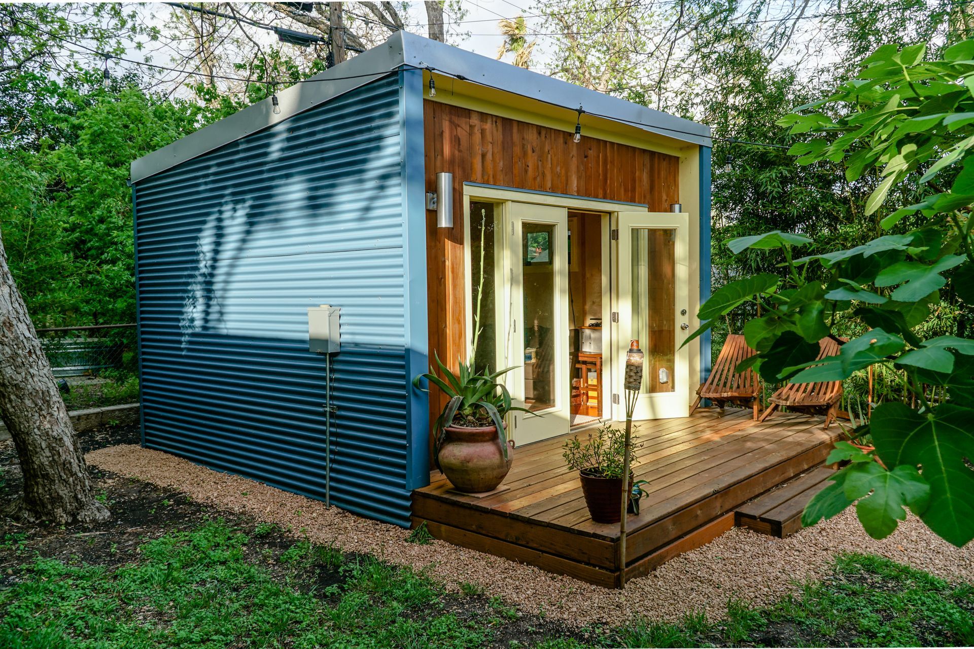Small, modern cabin with blue corrugated metal siding, wooden deck, and French doors, set in a yard with gravel and foliage.