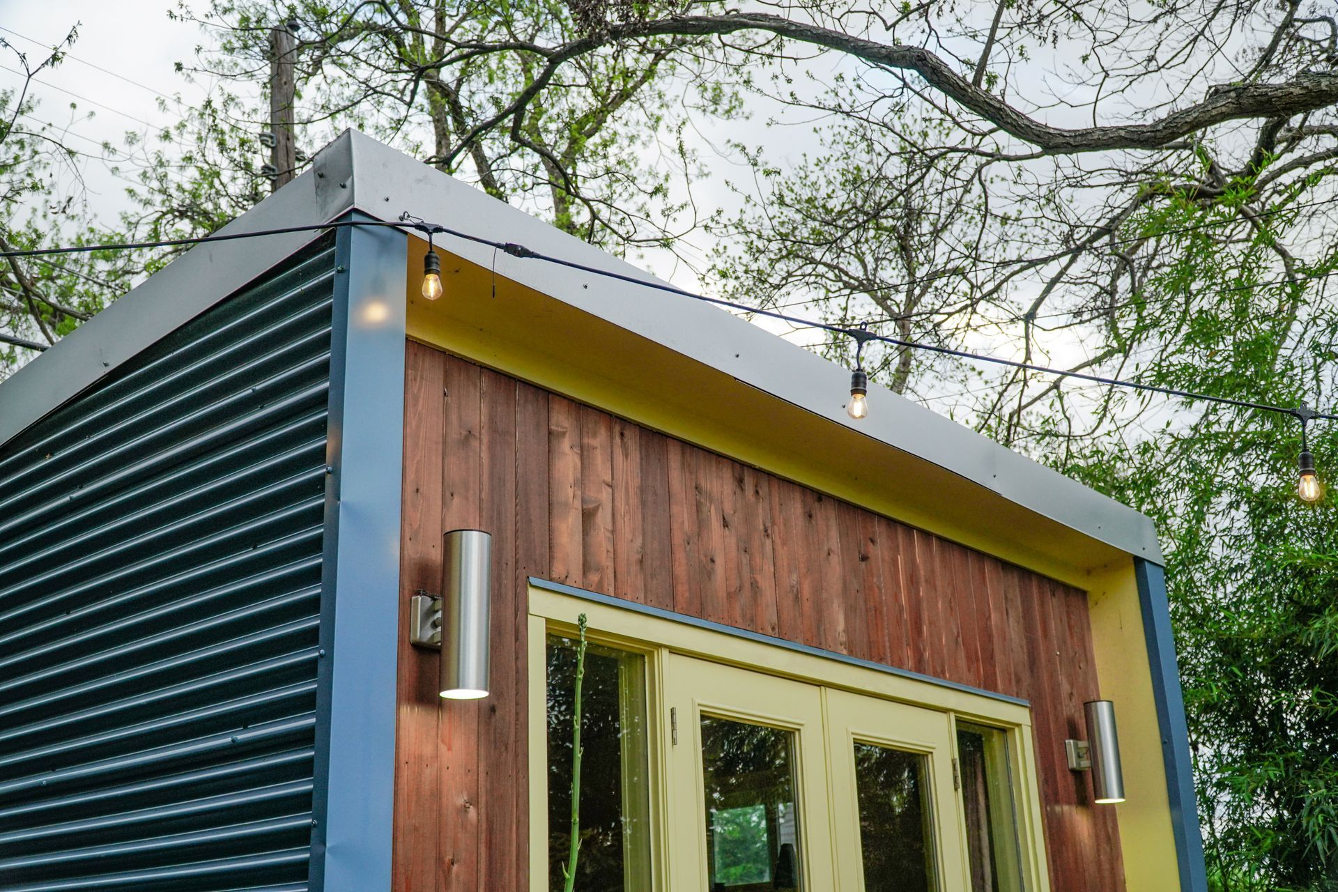 Modern outdoor shed with corrugated dark siding and wooden paneling, string lights, and a yellow door.