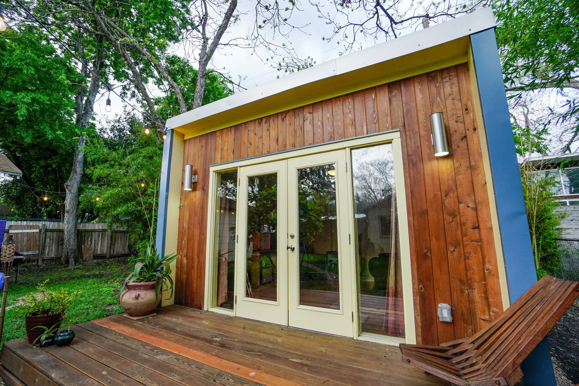 Wooden backyard shed with double glass doors, a deck, and surrounding greenery.