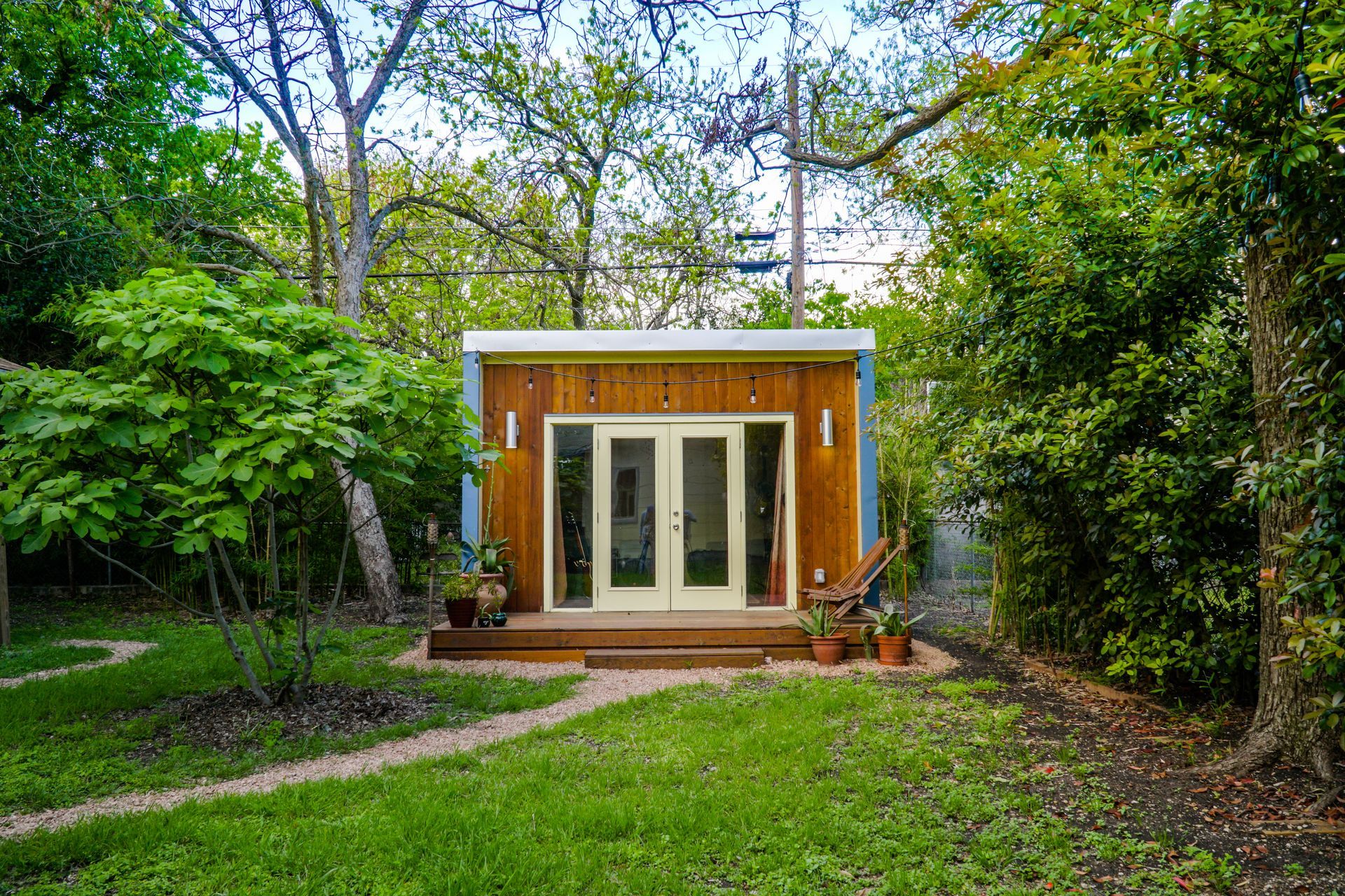 Wooden shed with double doors, on a small wooden deck, nestled in a backyard with trees and a grassy path.