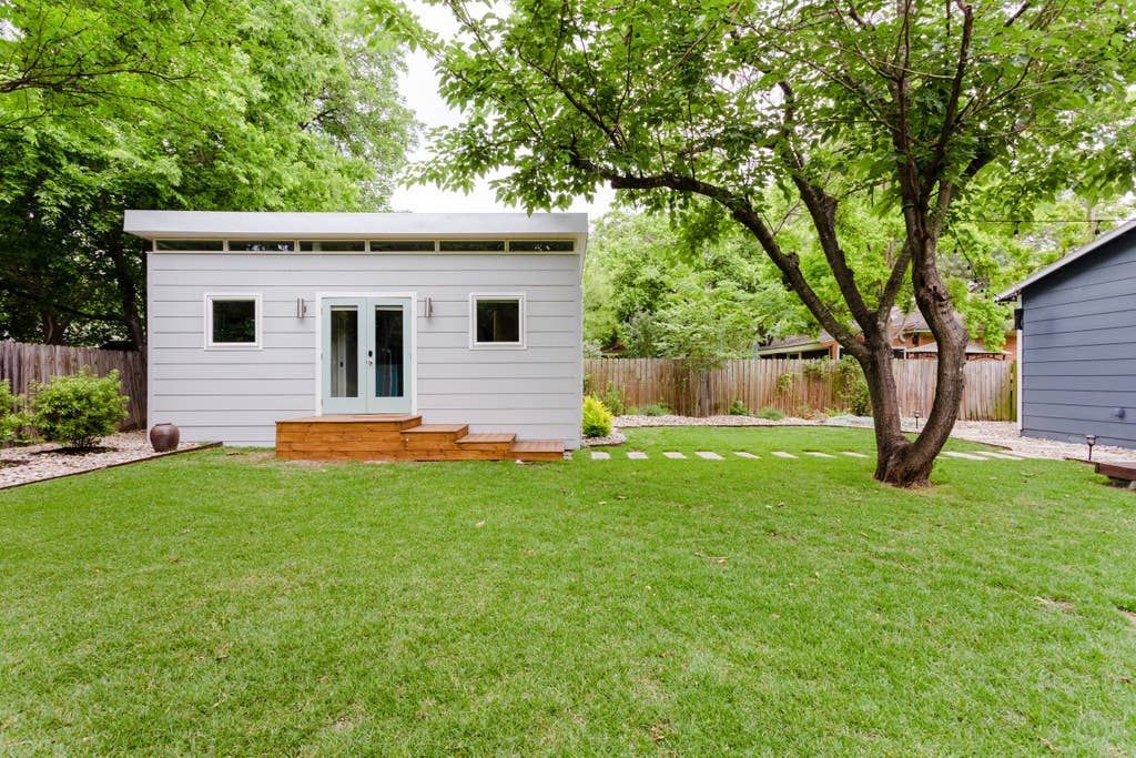 Small light grey backyard studio with a wooden deck and grass lawn.