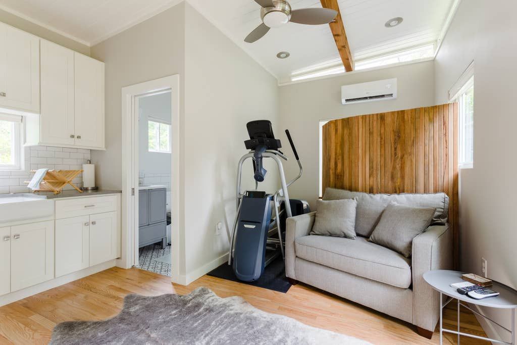 Small, bright room with workout equipment, sofa, and kitchen cabinets. Wooden floor and a decorative wood screen.