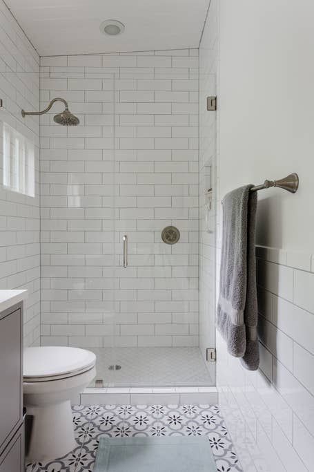 Small, white-tiled bathroom with glass shower, patterned floor, and a towel on a wall-mounted rack.