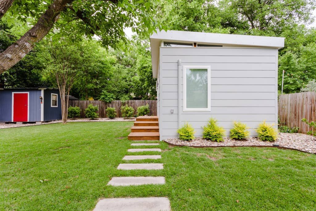 Stone path leads to a modern, light gray shed in a backyard with a red and blue shed in the background.