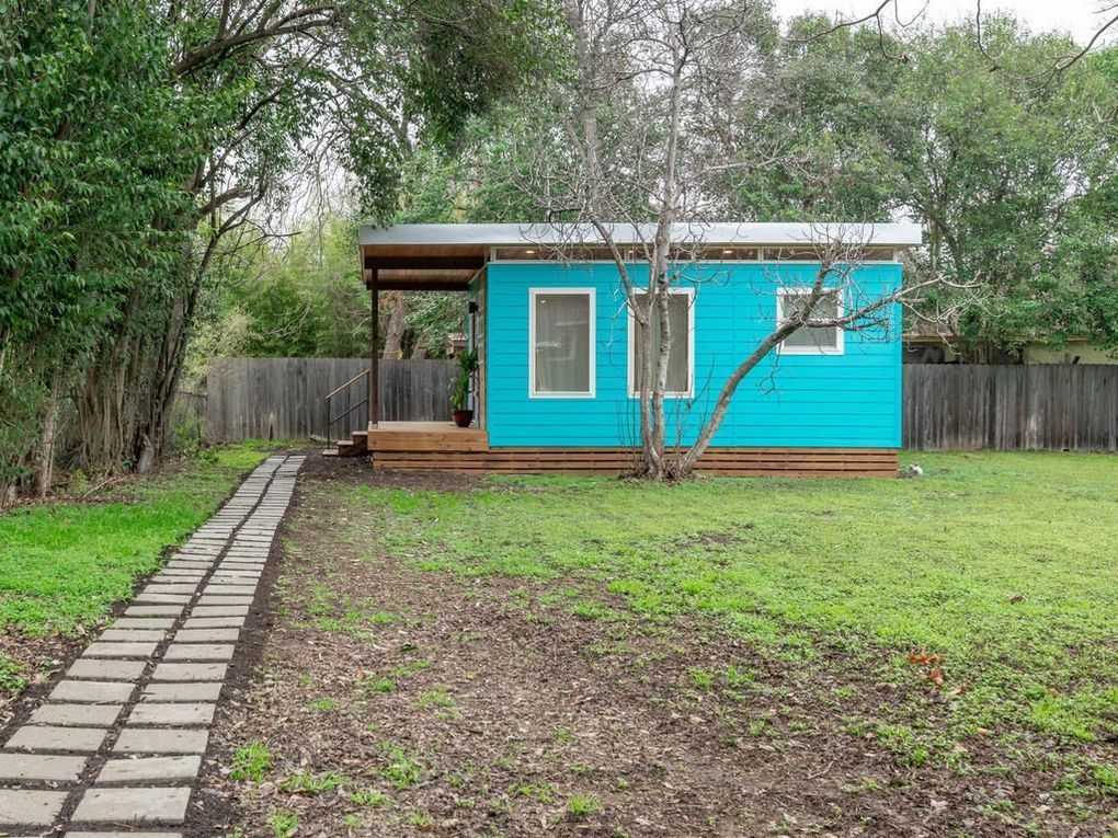 Bright blue tiny house with a covered porch, set in a backyard with a stone path.