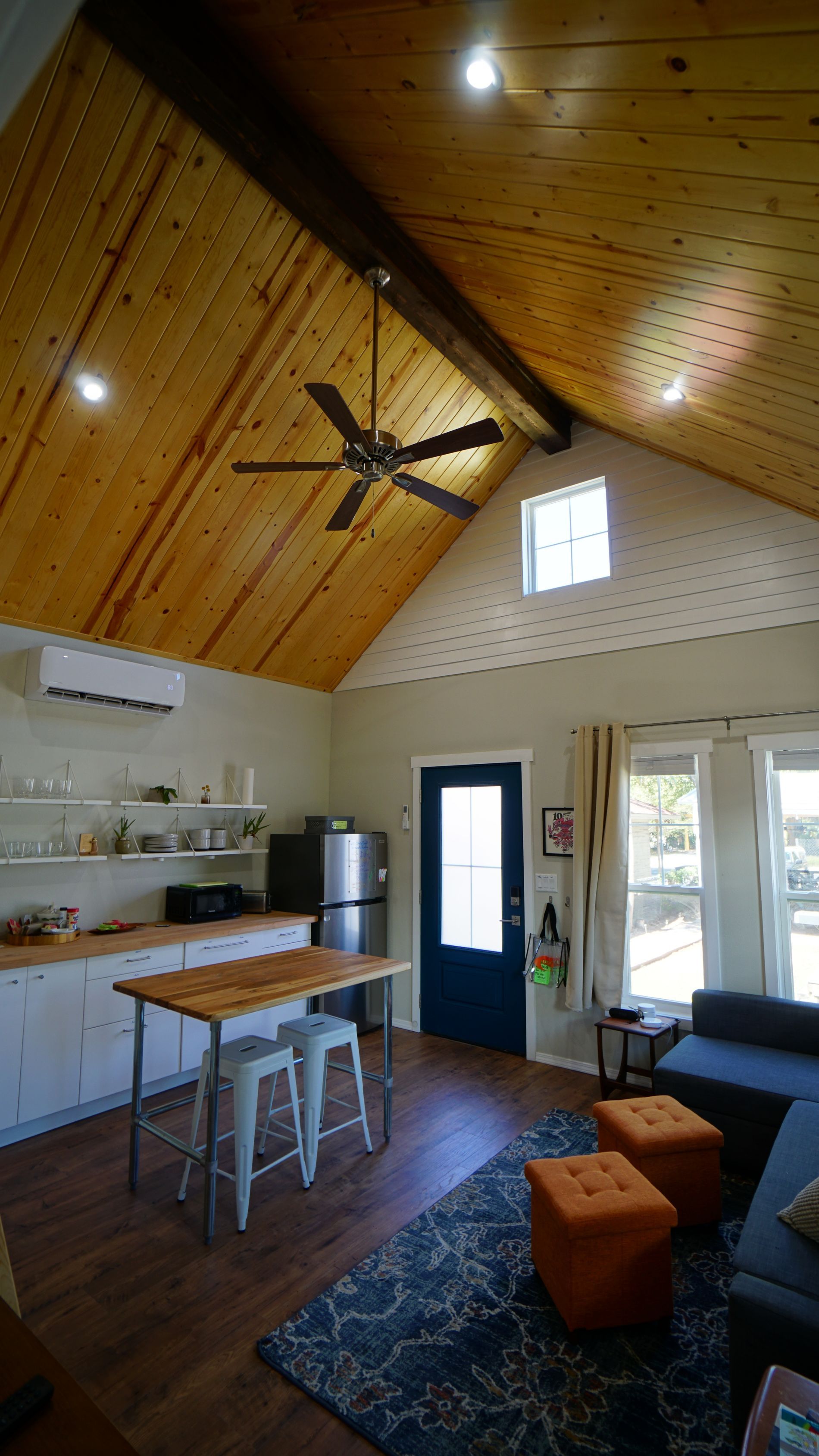 Interior view of a cabin with wood ceiling, kitchen, blue couch, rug, and ottomans.