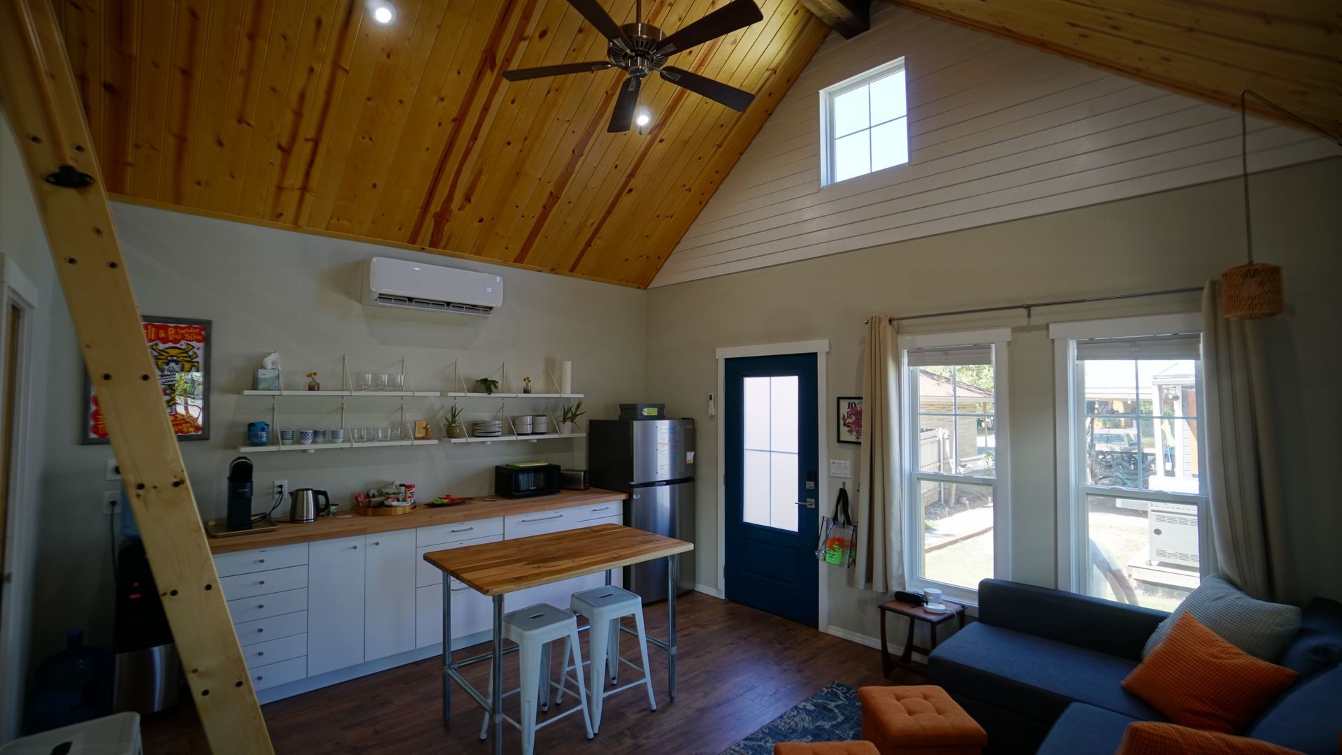 Small cabin interior with wood-paneled ceiling, kitchen area, living space with blue sofa, and small loft access.