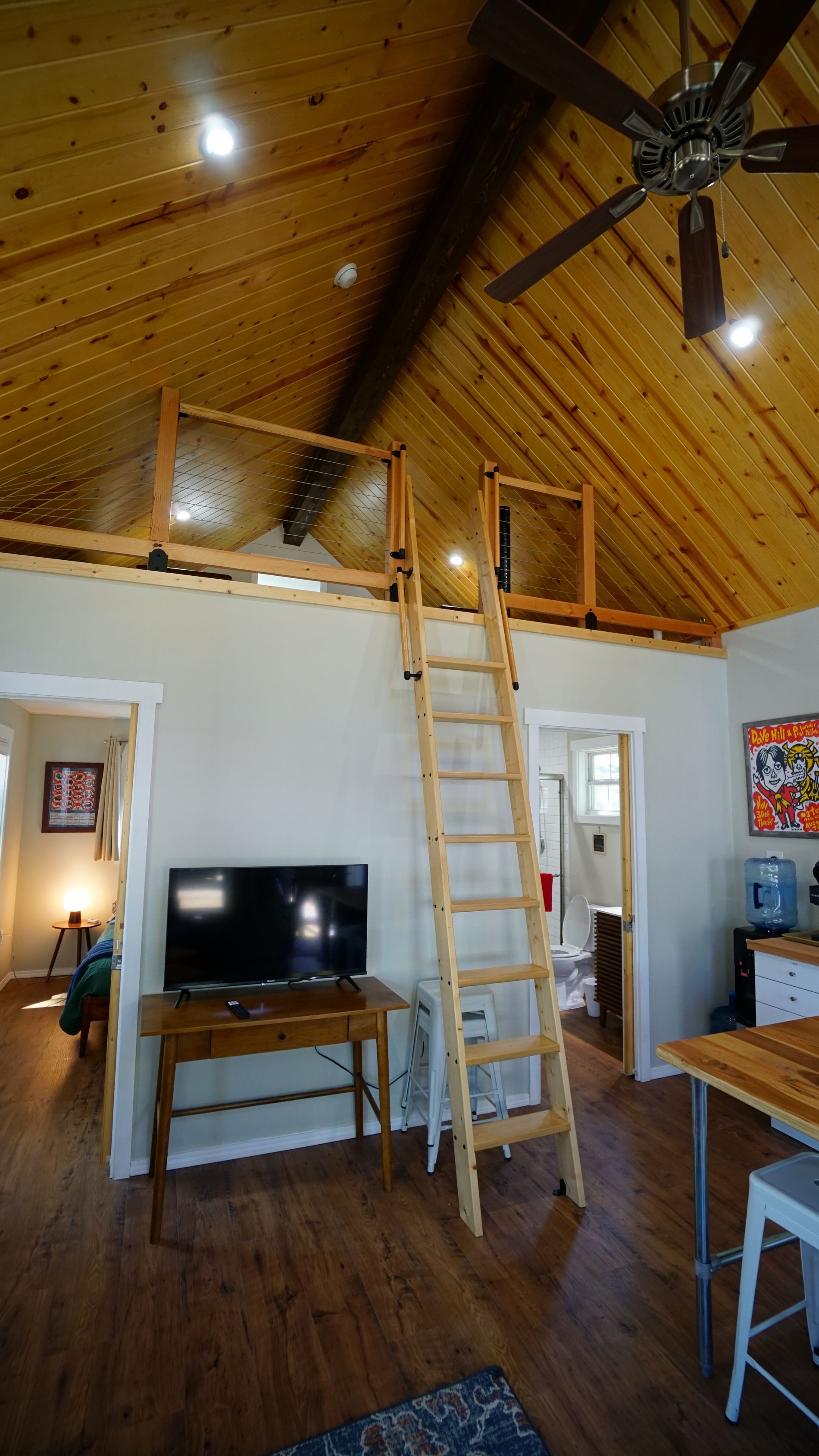 Interior of a cabin with a loft, wooden ceiling, ladder, TV, and open doorways.
