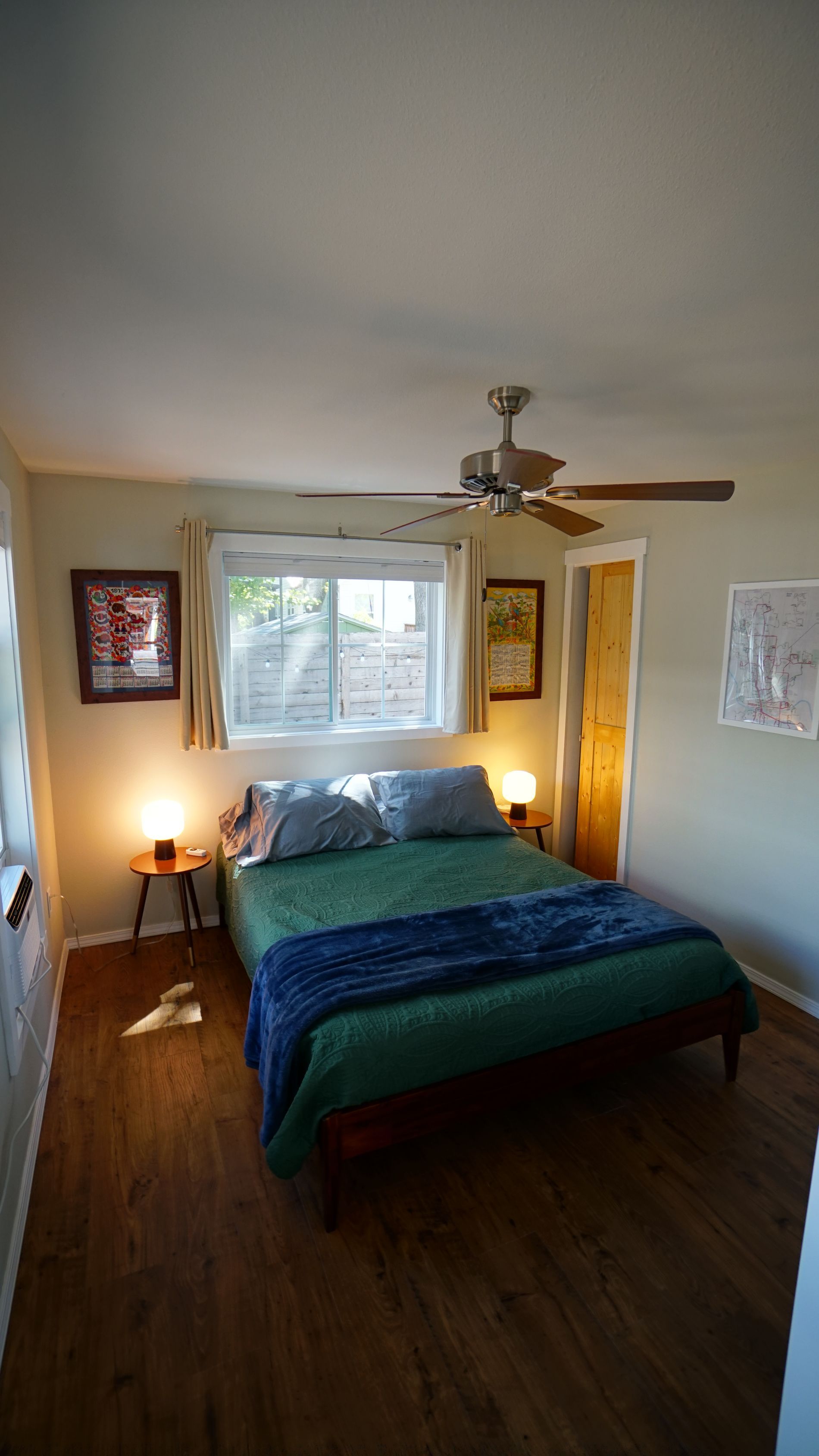 Bedroom with bed, nightstands, window, artwork, and ceiling fan. Green bedding with blue throw, wood floors.