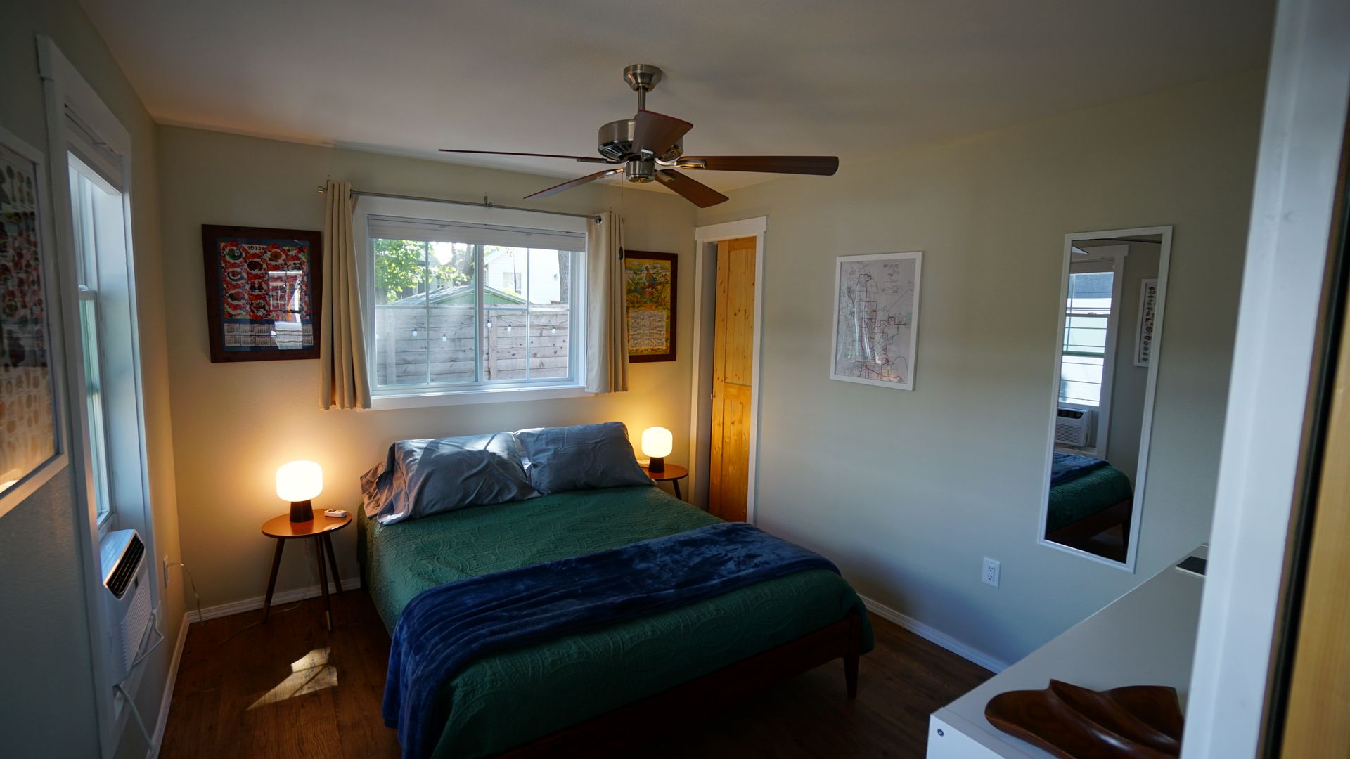 Small bedroom with a bed, window, and mirror. Green bedding, wooden floors, and a ceiling fan.