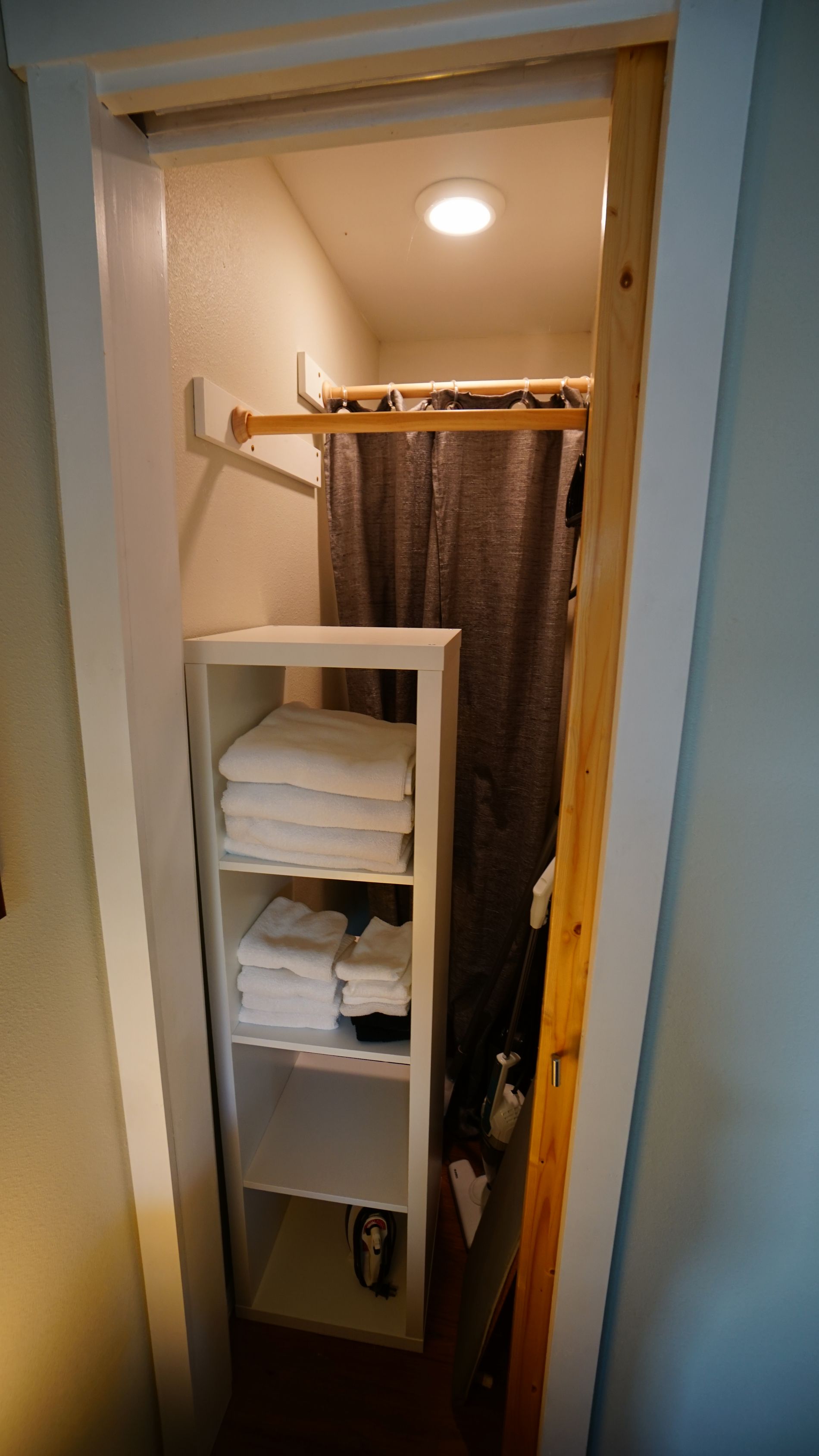 A small closet with white shelves holding towels. A wooden rod hangs above a gray curtain.