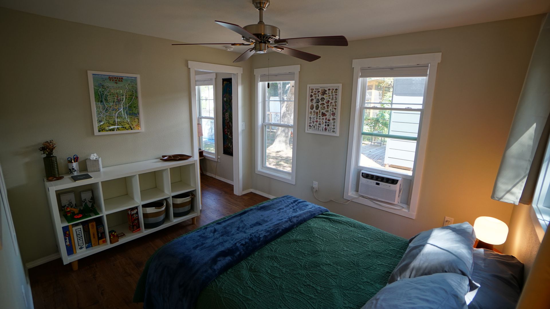 Bedroom with bed, windows, bookcase, and ceiling fan. Green and blue bedding, cream walls, and hardwood floors.