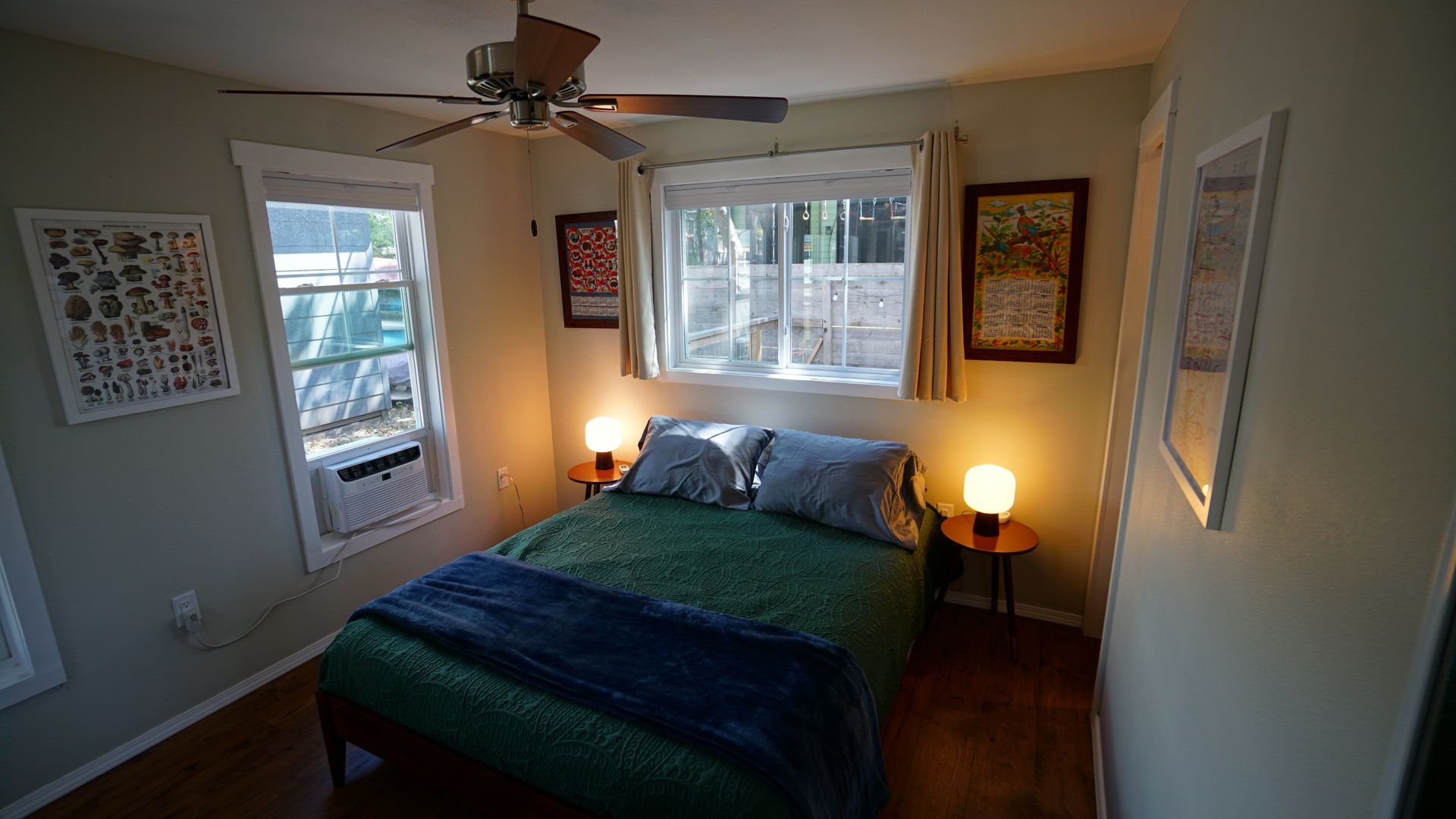 Cozy bedroom with a bed, small windows, two bedside tables with lamps, and artwork on the walls.