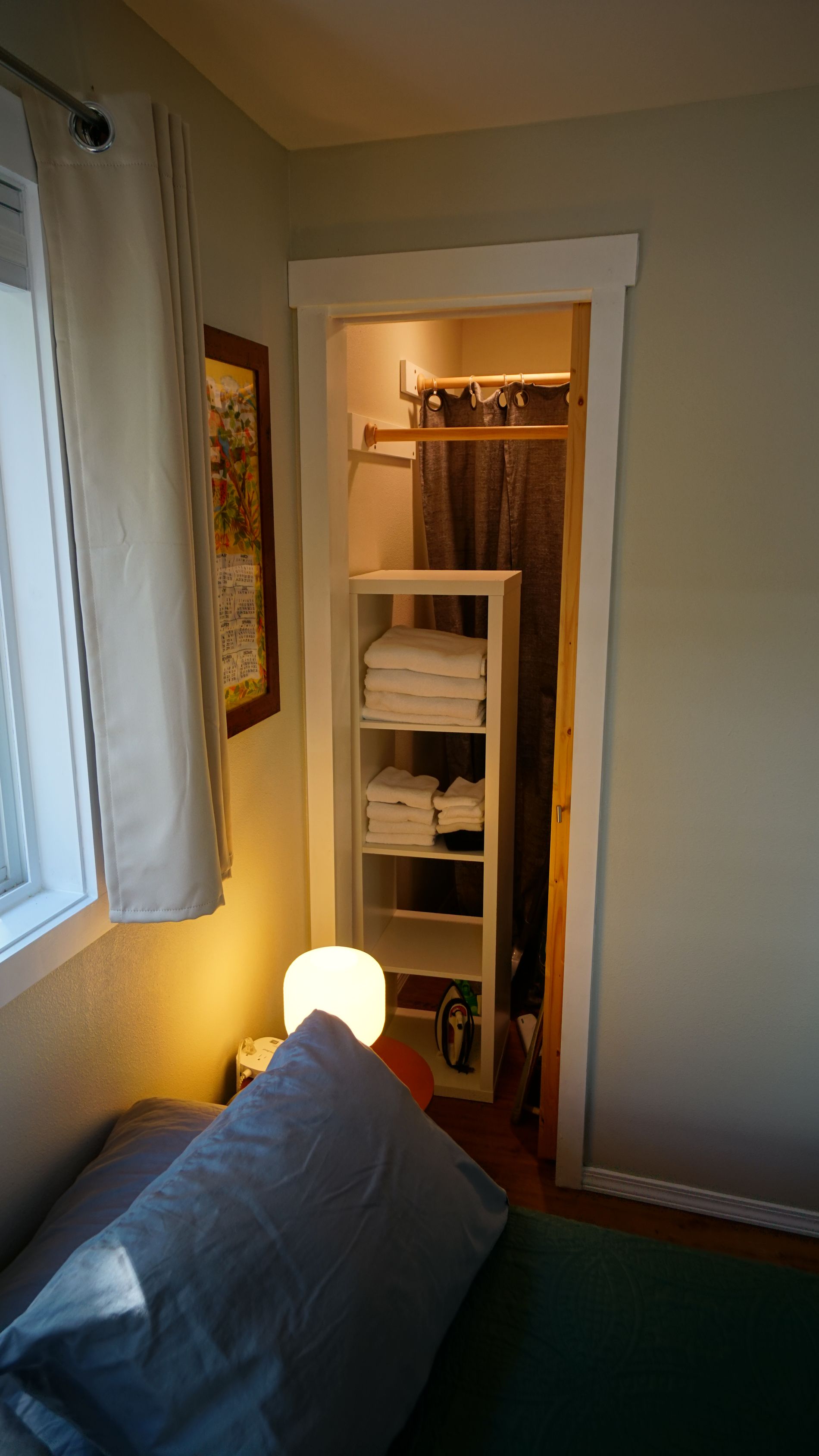 Bedroom closet with white shelving, rod, and door frame. Light on, bed in foreground.