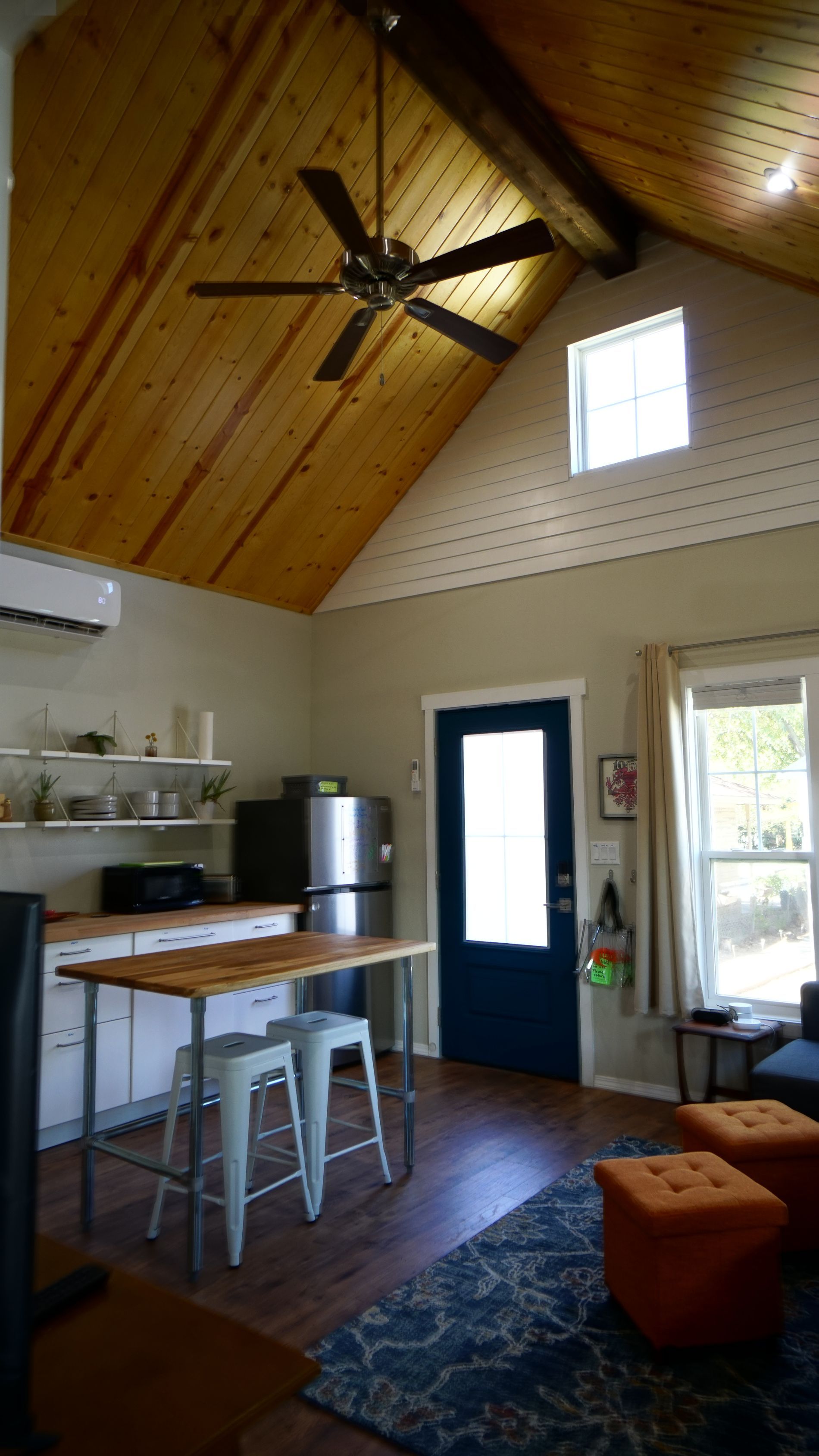Small living space with a wood-paneled ceiling, dark blue door, kitchen, and seating area with orange ottomans.