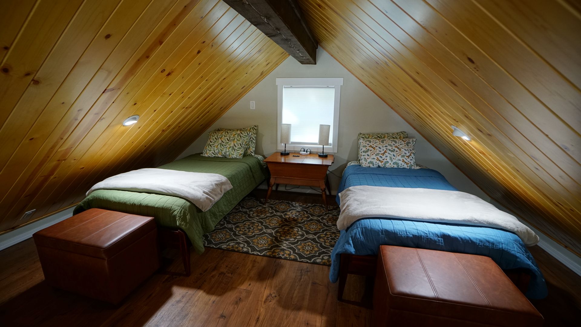 Attic bedroom with two twin beds, small window, wooden ceiling, and dark brown ottomans.