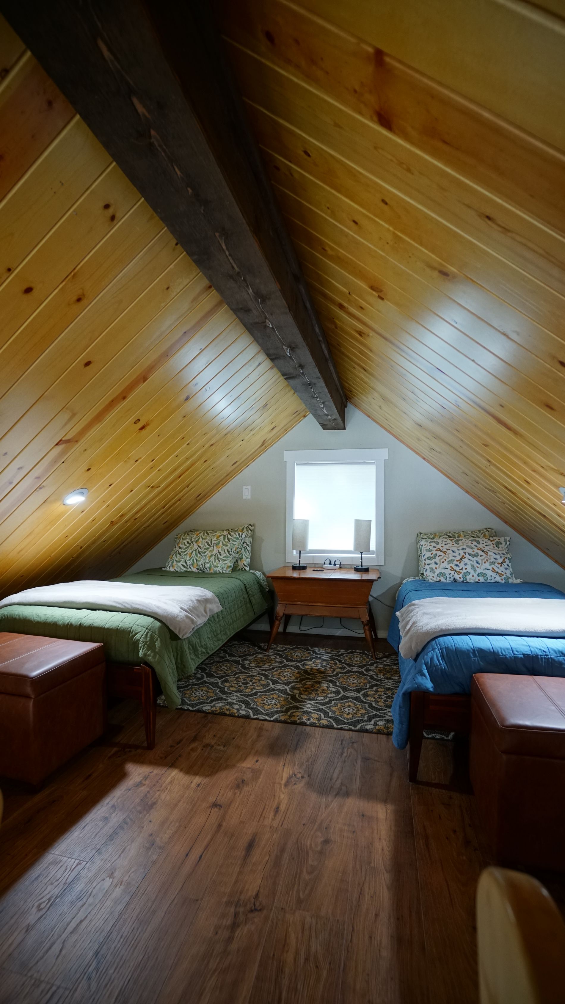 Bedroom with two beds under sloped wooden ceiling. A window is between the beds.