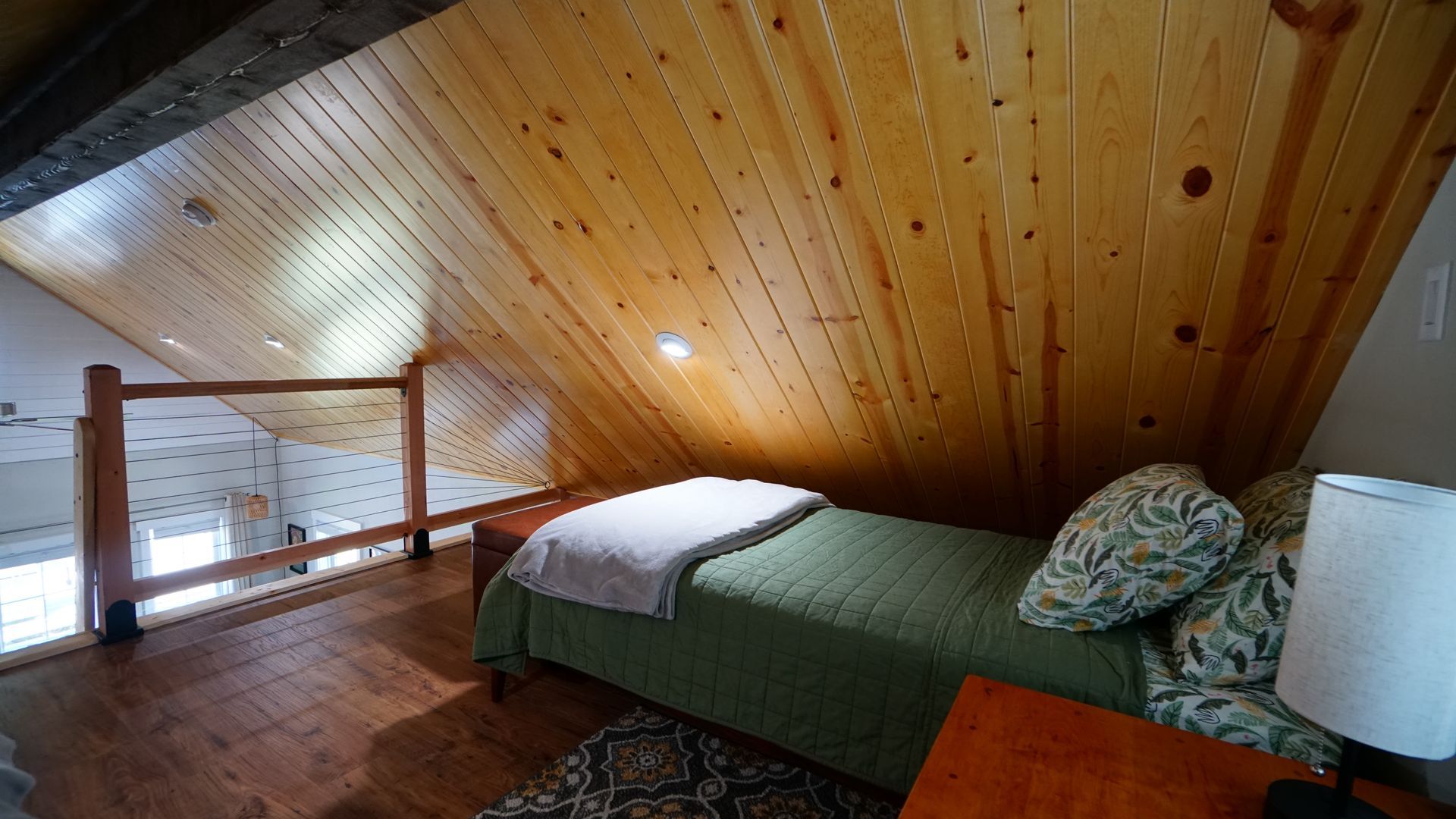 Loft bedroom with wooden walls and flooring. Bed with green quilt and pillows, small bedside table and lamp.
