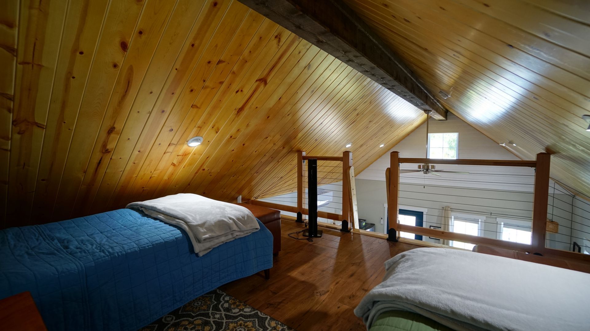 Loft bedroom with wooden walls and two beds: one blue, one white. View of the lower level is in the background.