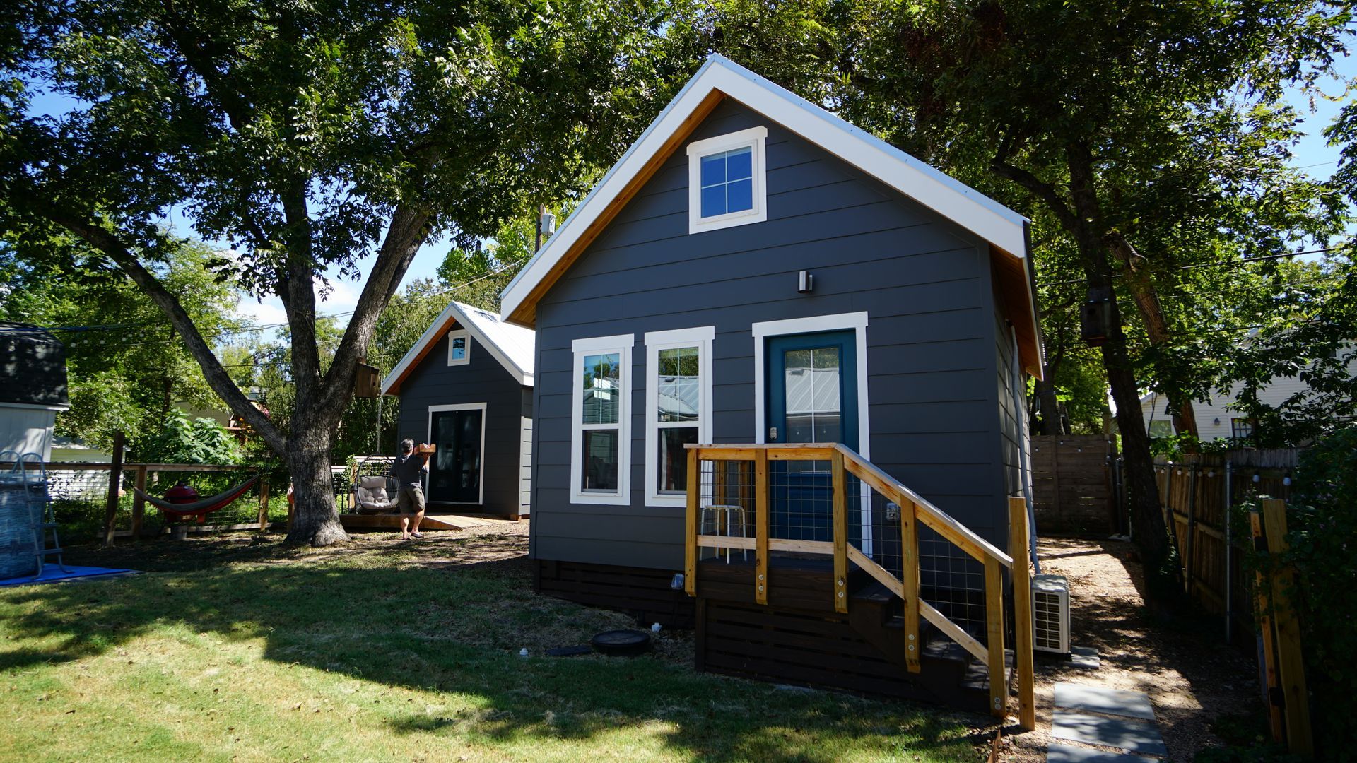 Two small, blue-gray houses with white trim and wooden steps, surrounded by trees and a grassy yard.