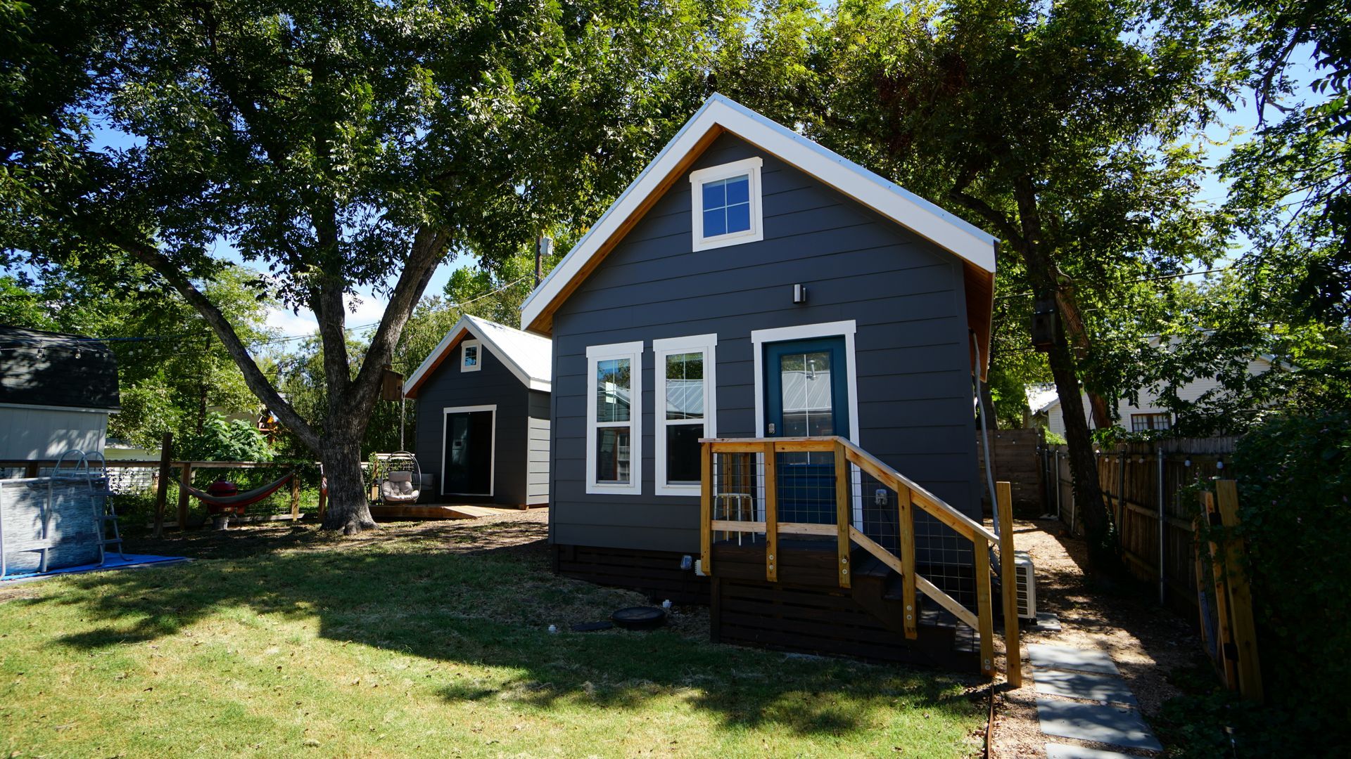 Small dark blue house with white trim, front porch, and small backyard building, sunny day.