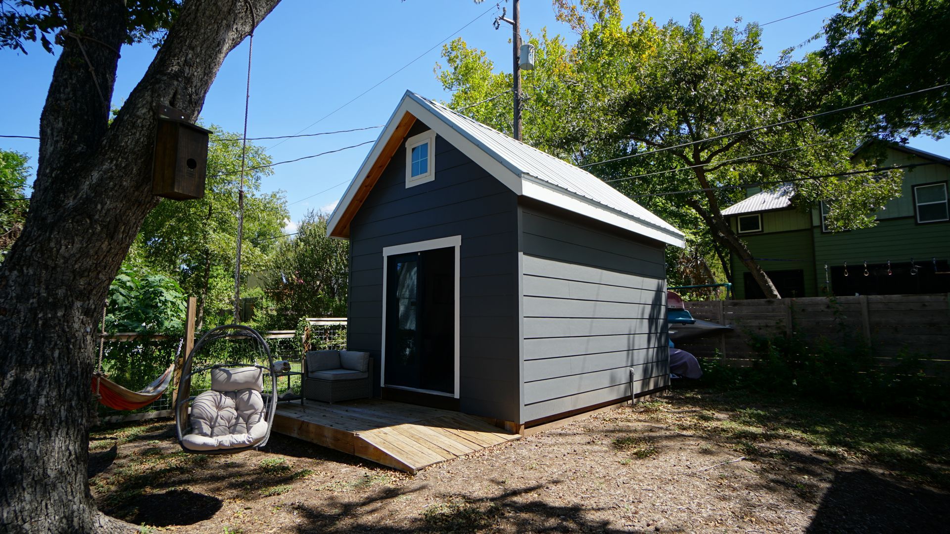 A small, gray shed with a ramp and a birdhouse in a yard with a swing and trees.