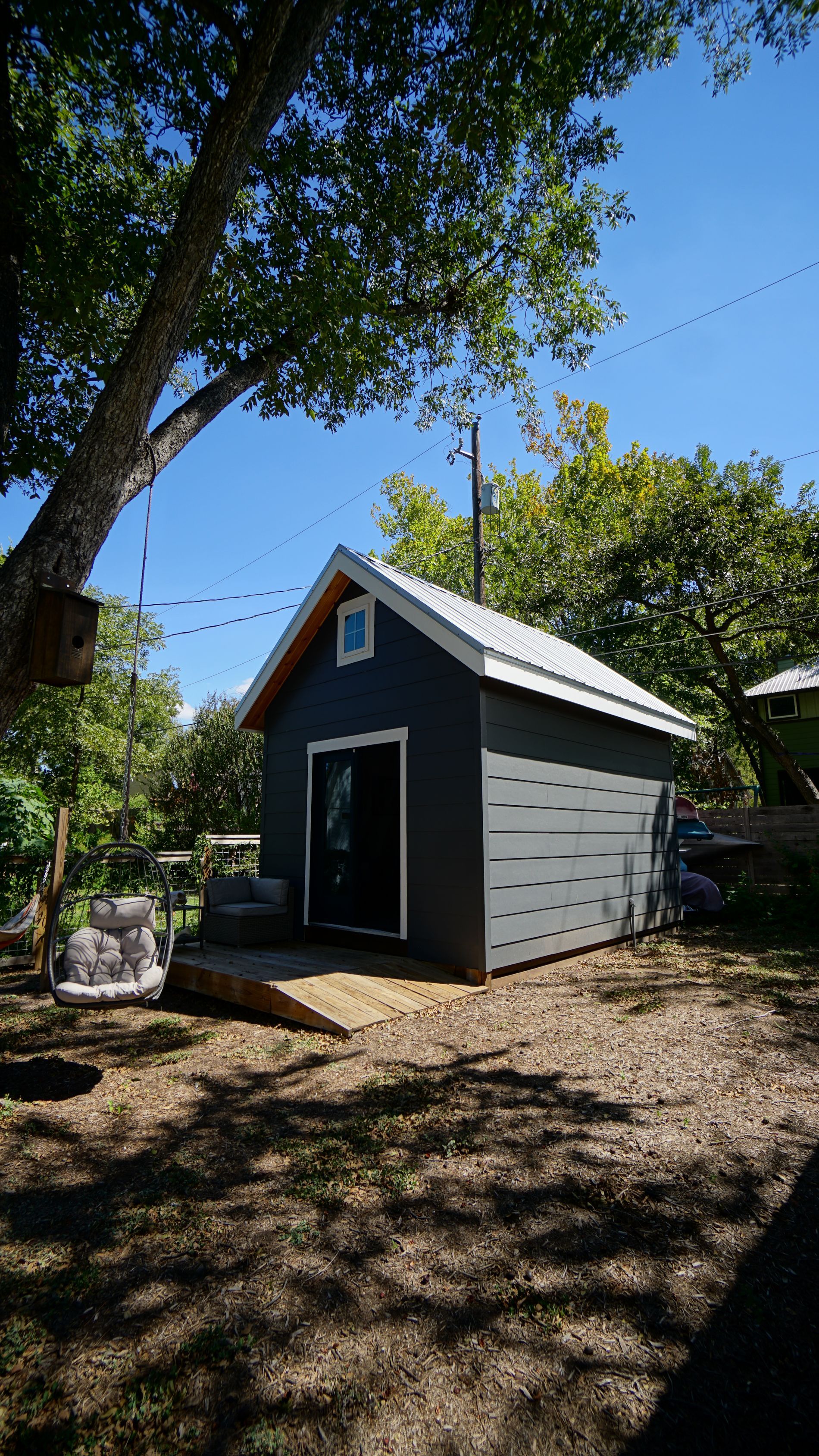 Small gray shed with white roof and a small wooden deck outdoors, under trees.