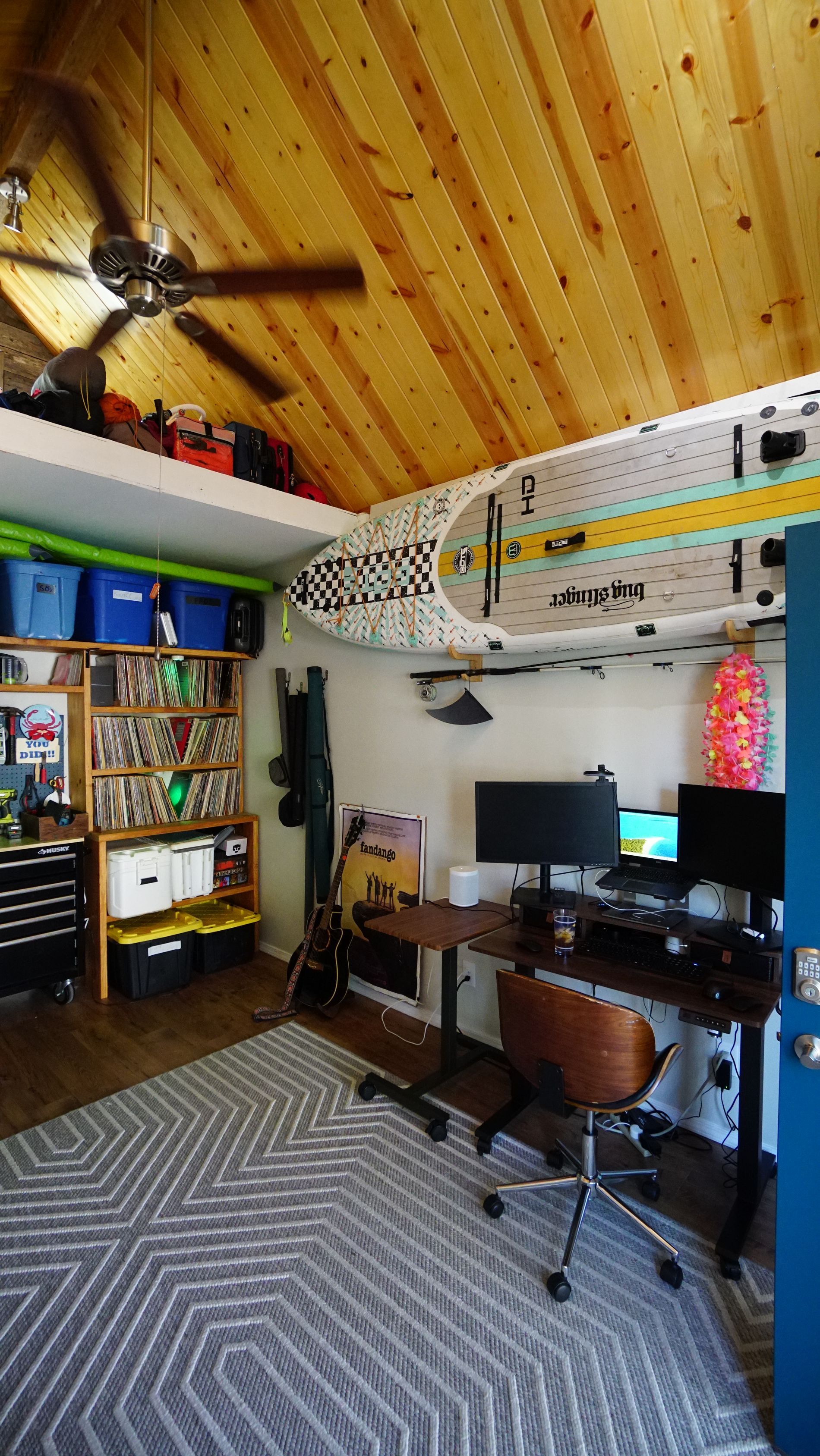 Room with wooden ceiling, surfboard, desk with two monitors, shelving, and a patterned rug.