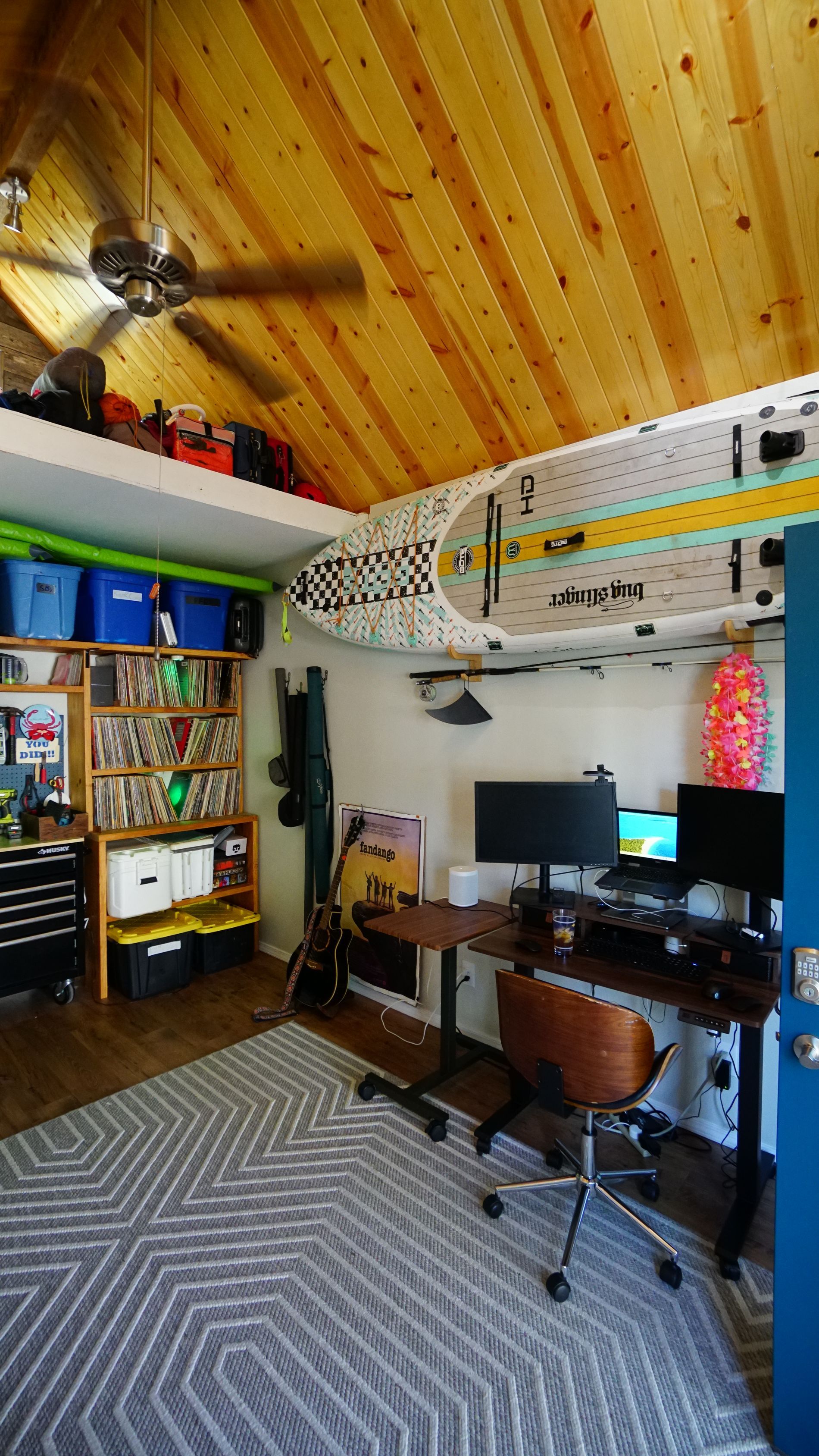Home office with wooden ceiling, surfboard, desk, shelving, and rug.