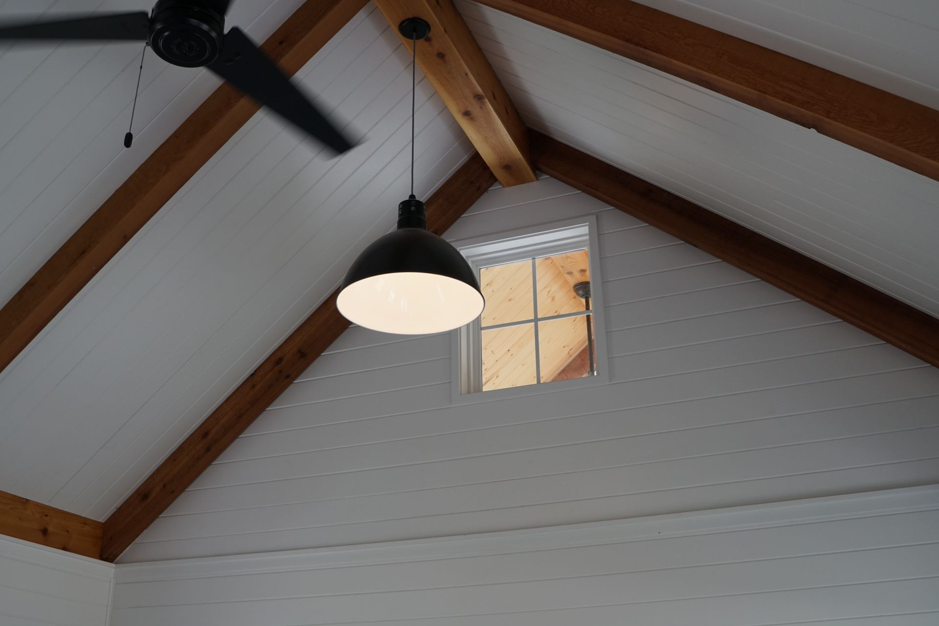 Inside view of a white vaulted ceiling with wooden beams, a black pendant light, and a small window.