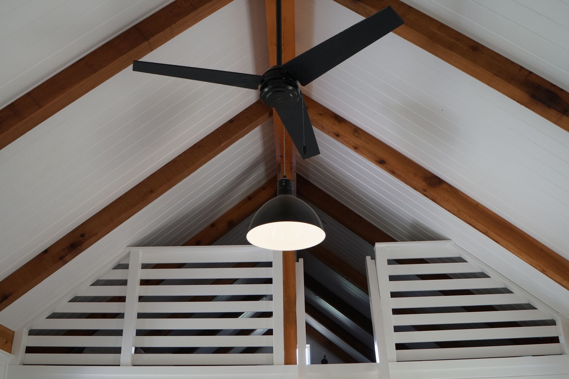 Black ceiling fan and pendant light hanging from a white sloped ceiling with brown beams, over white shuttered windows.