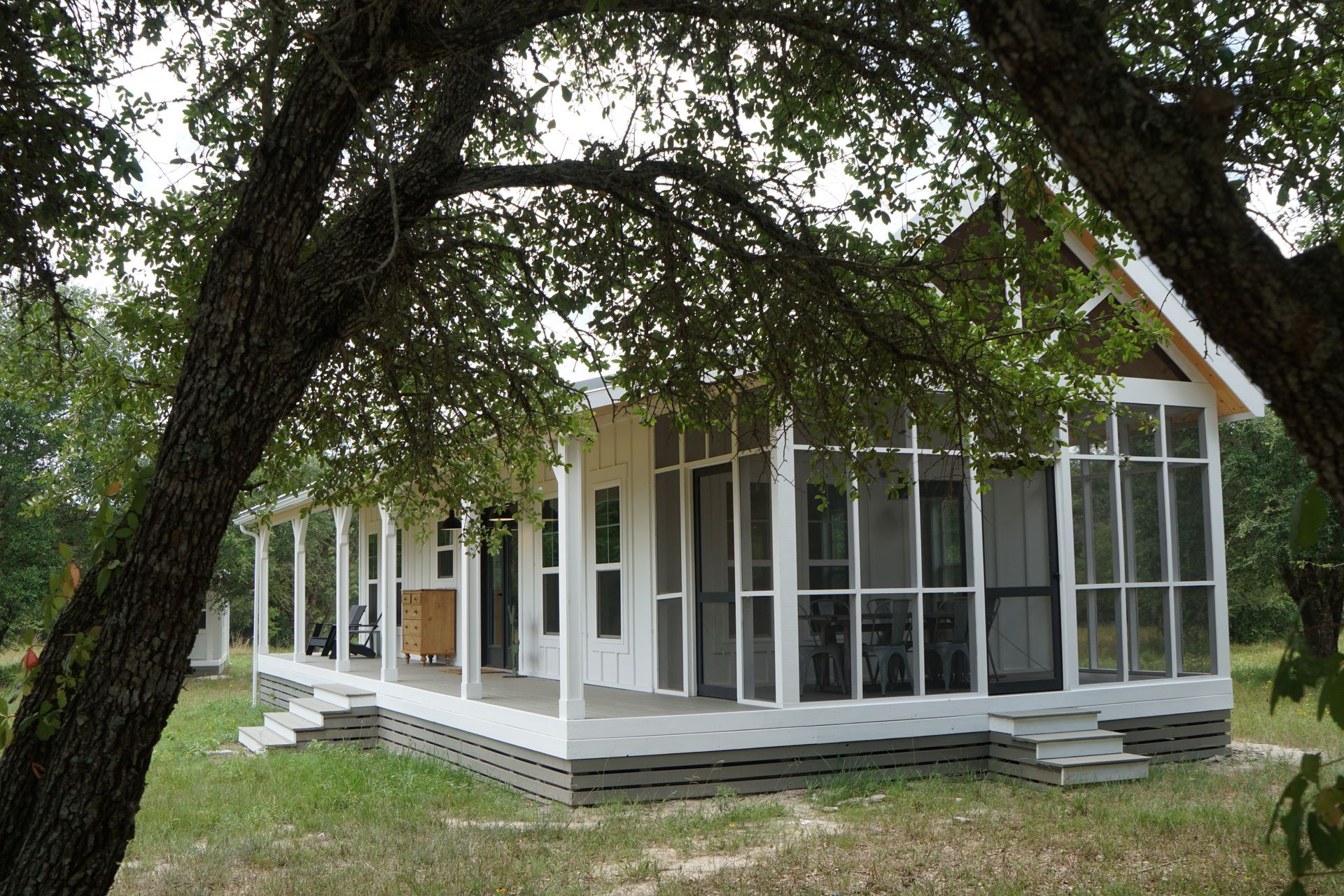 White cottage with porch and screened-in area, framed by tree branches in a grassy field.