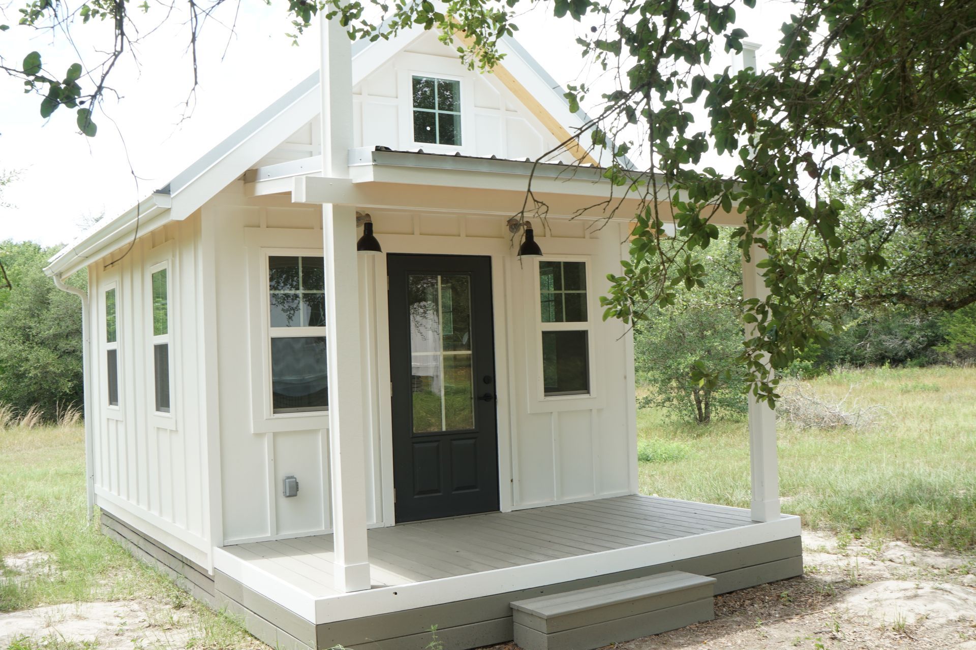 White tiny house with a black door, small porch, and gabled roof, set in a grassy area.