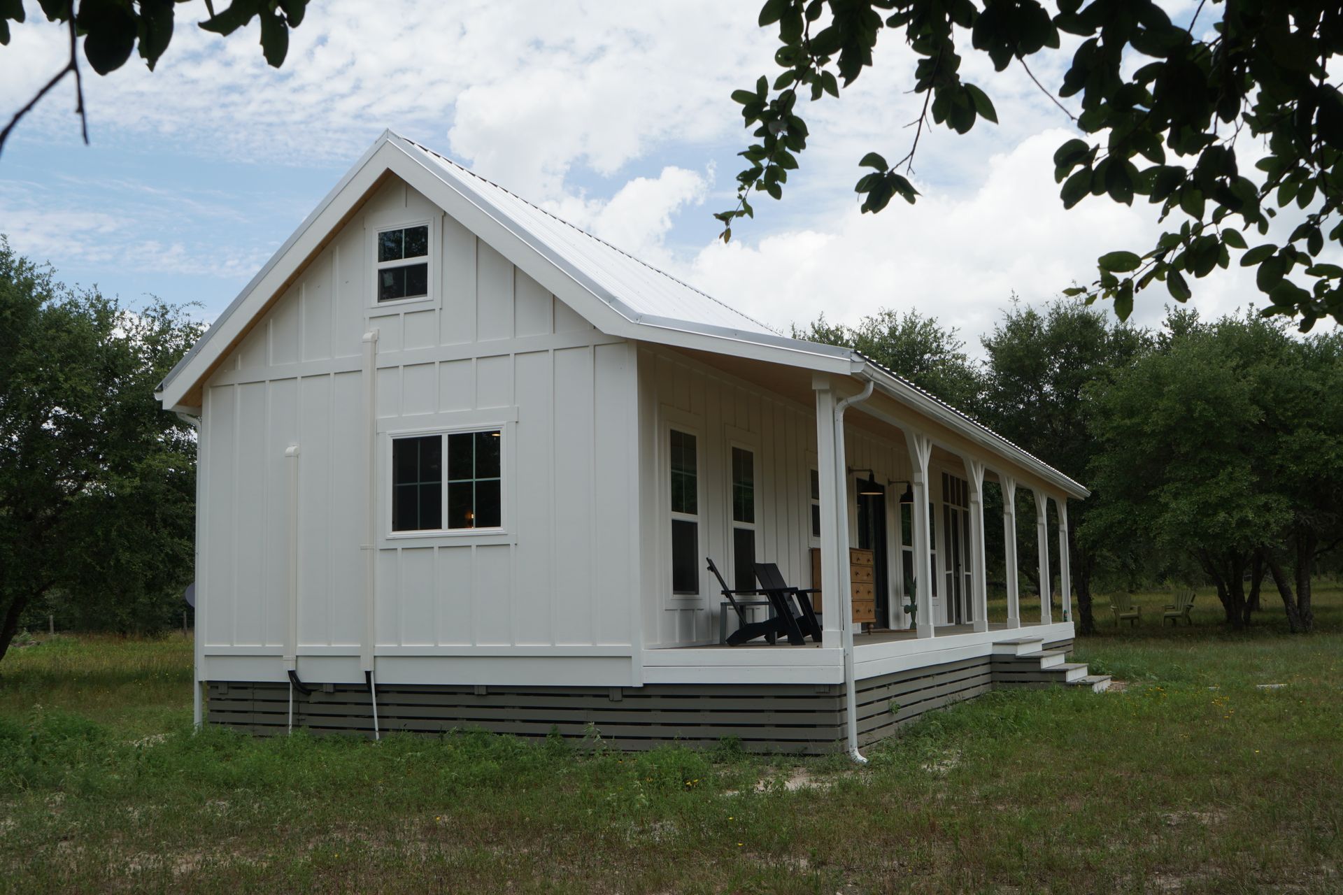 White cottage with porch and metal roof in a grassy field under a cloudy sky.