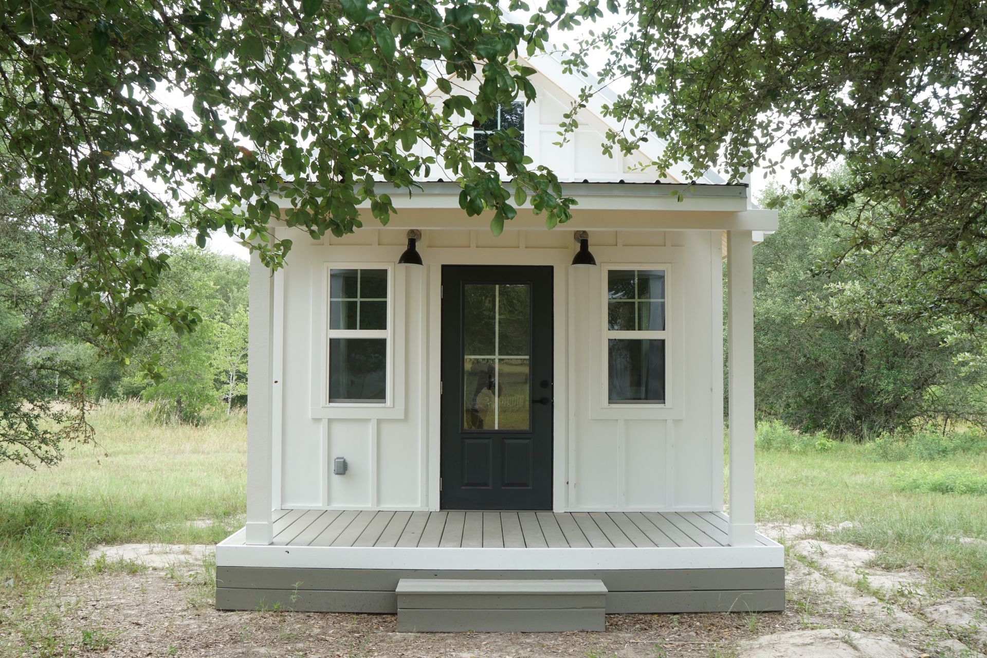 White tiny house with black door and porch, surrounded by greenery.