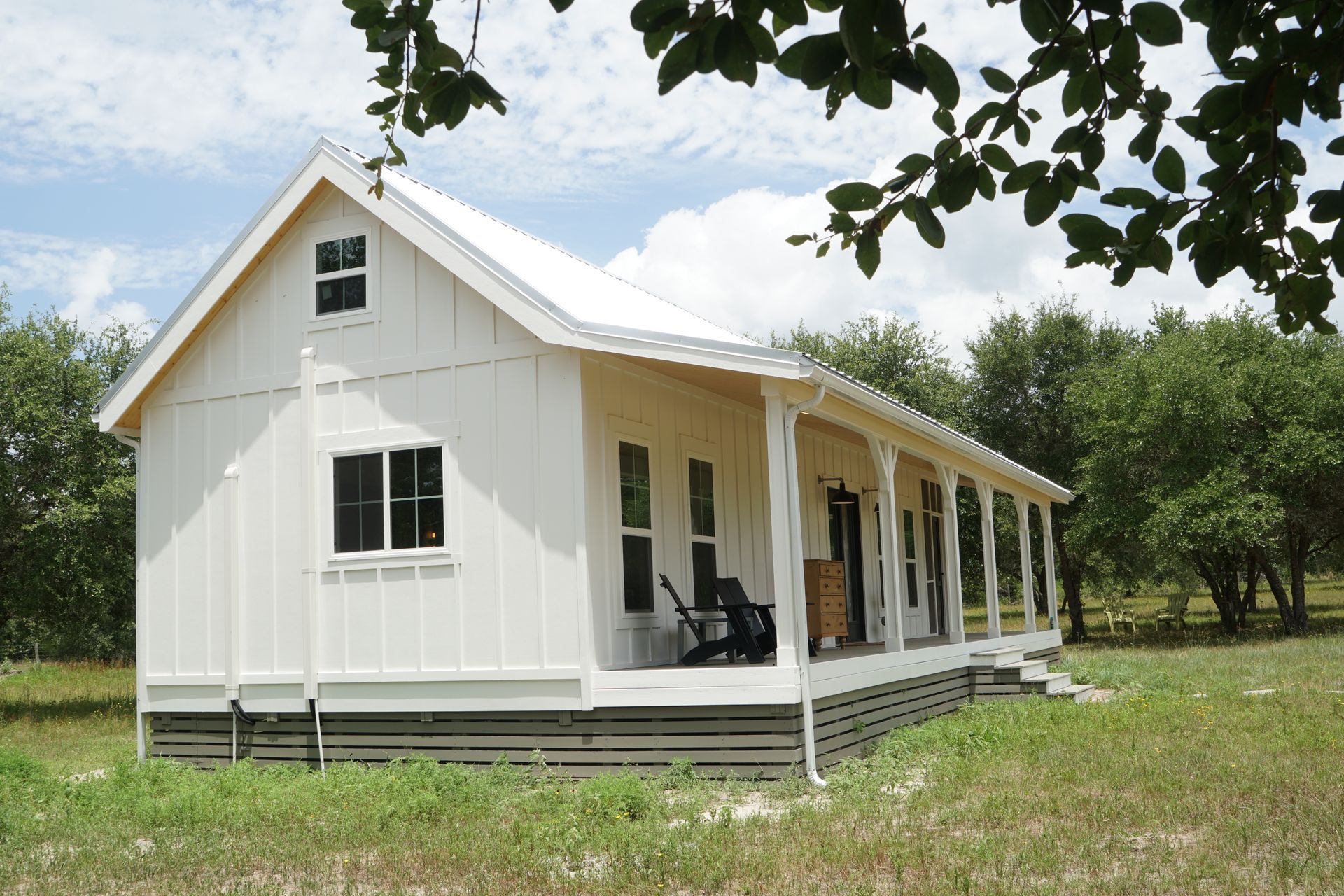 White cottage with a porch, surrounded by trees and tall grass, under a bright sky.
