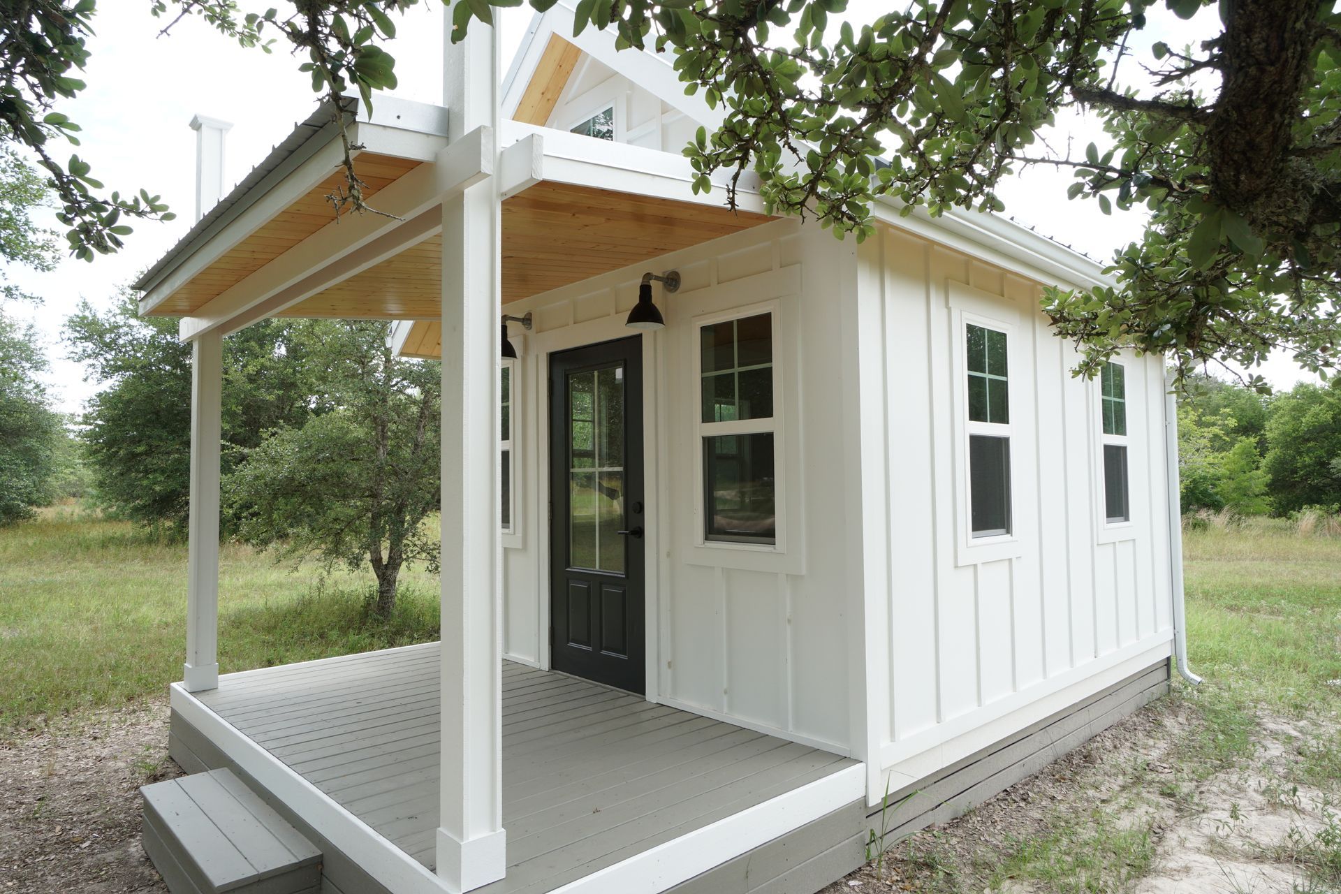 White tiny house with porch and black door; set in a grassy field with trees.