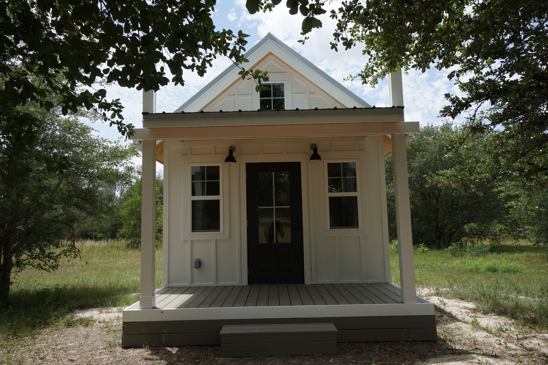 Small white cabin with porch, black door, and two windows; set in a wooded area.