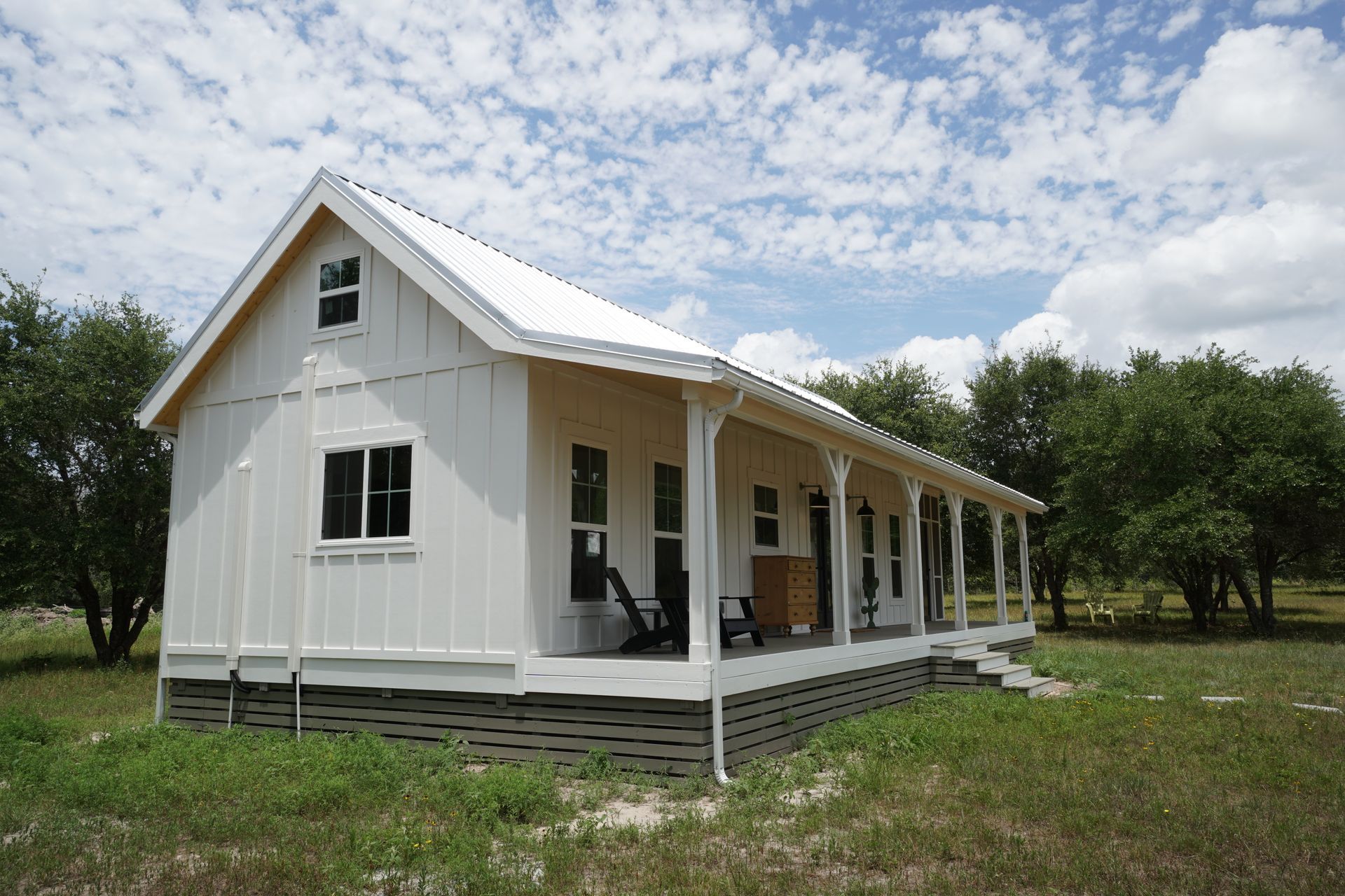 White farmhouse with a long porch, white roof, and two windows; set in a grassy yard under a cloudy sky.