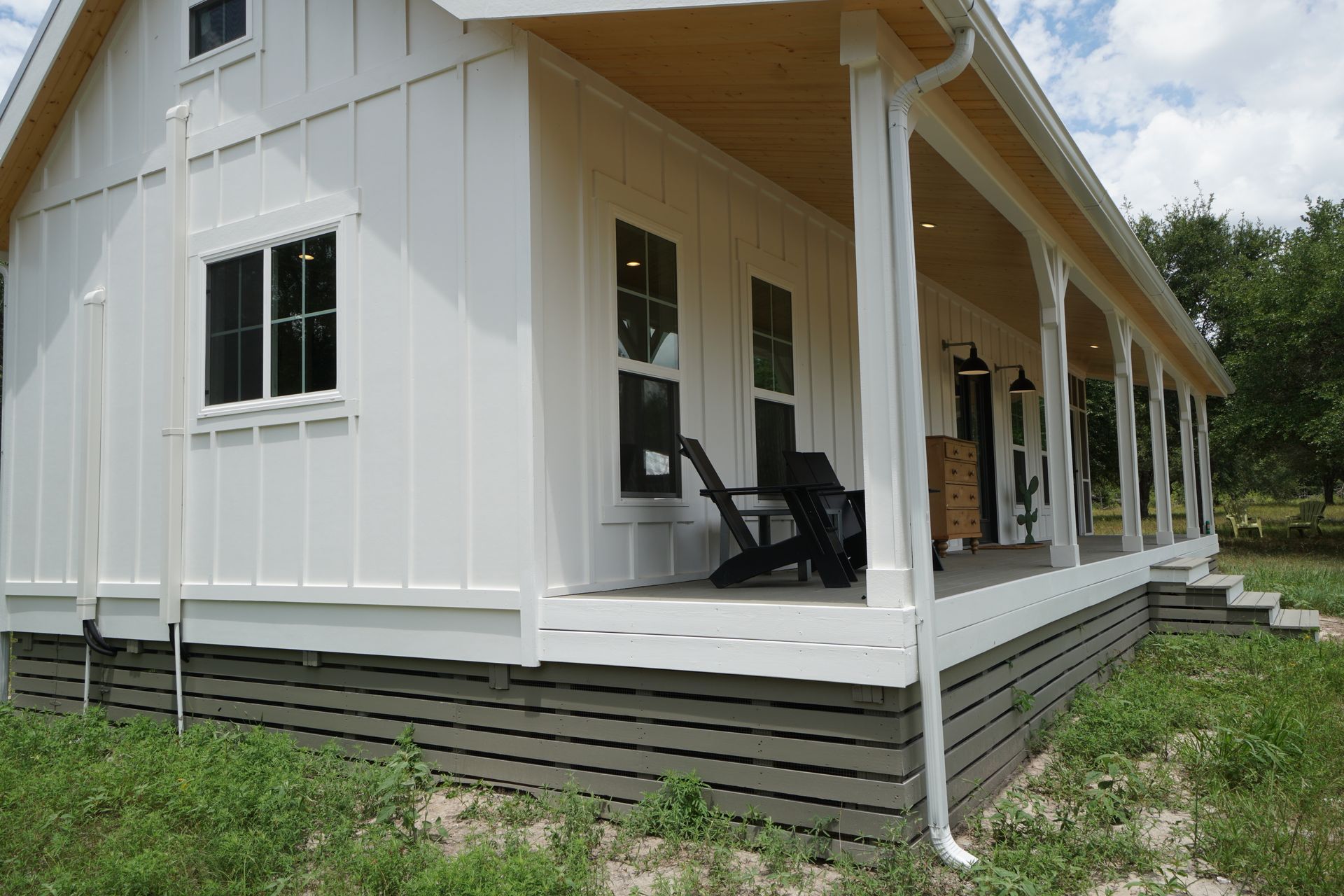 White farmhouse with a long porch, gray skirting, and black windows.