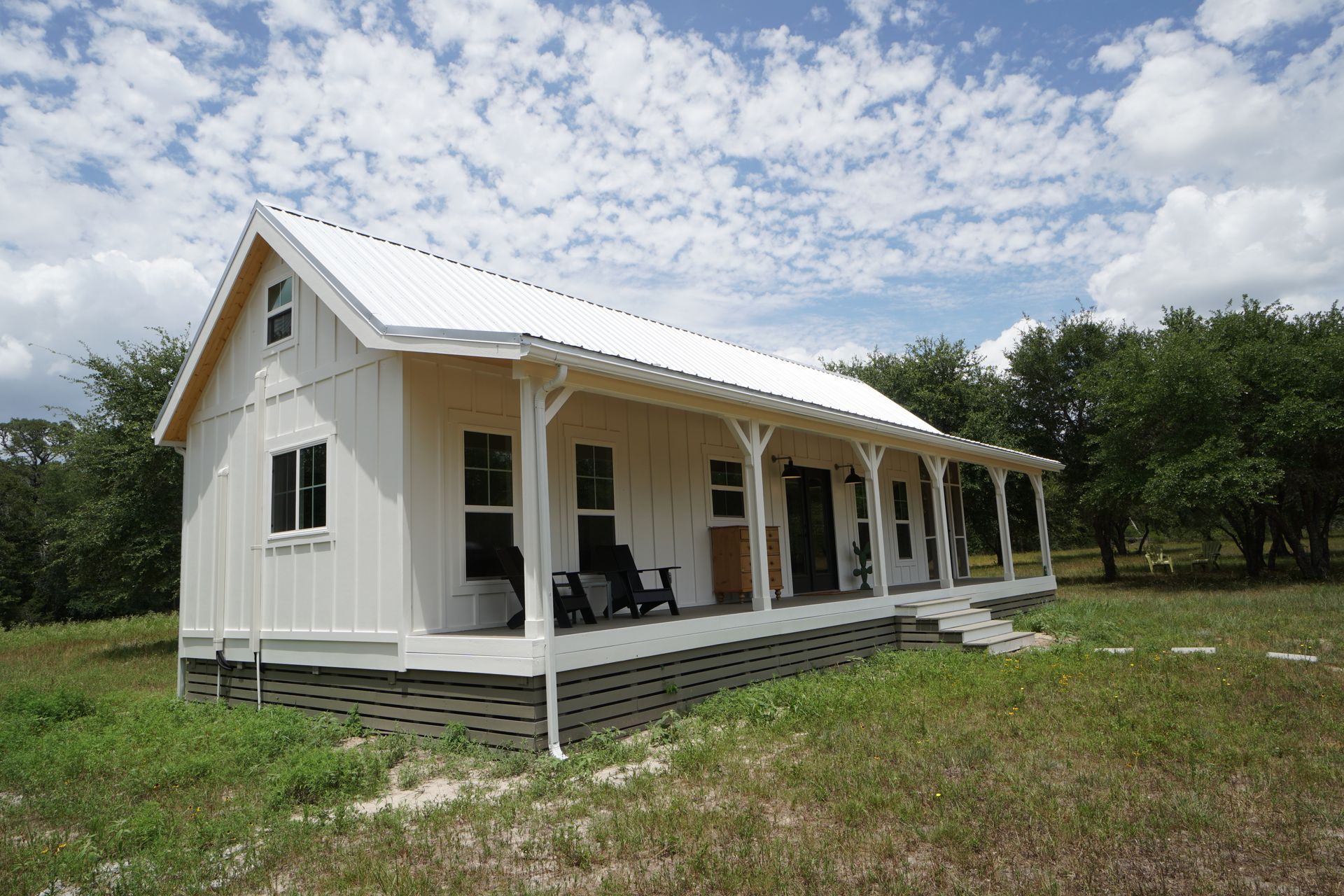 White farmhouse with porch and white metal roof in a grassy field under a cloudy sky.
