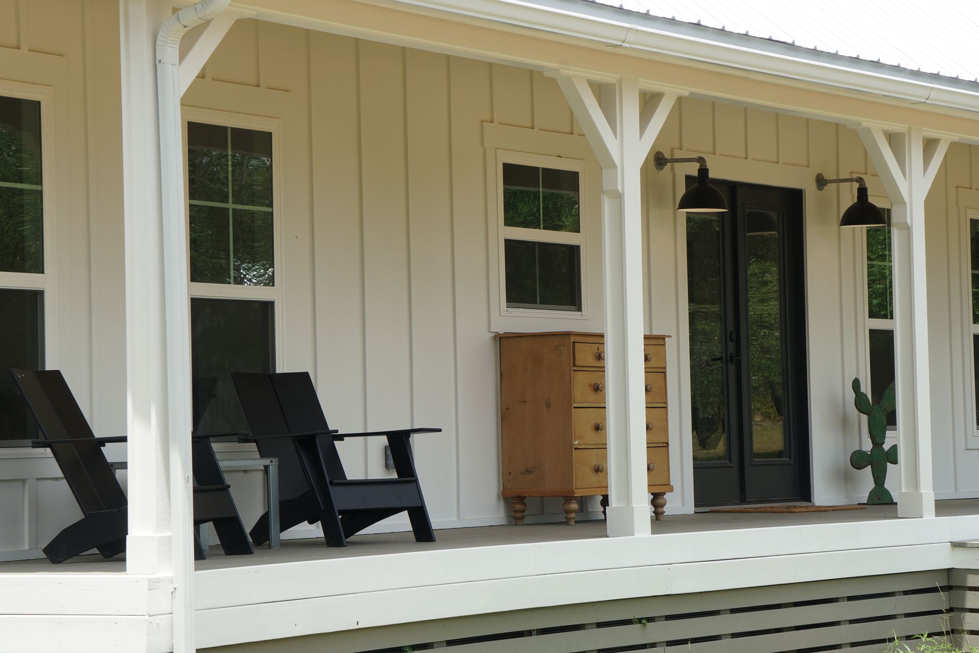 White farmhouse porch with black chairs, doors, and lights. A wooden chest and cactus complete the scene.