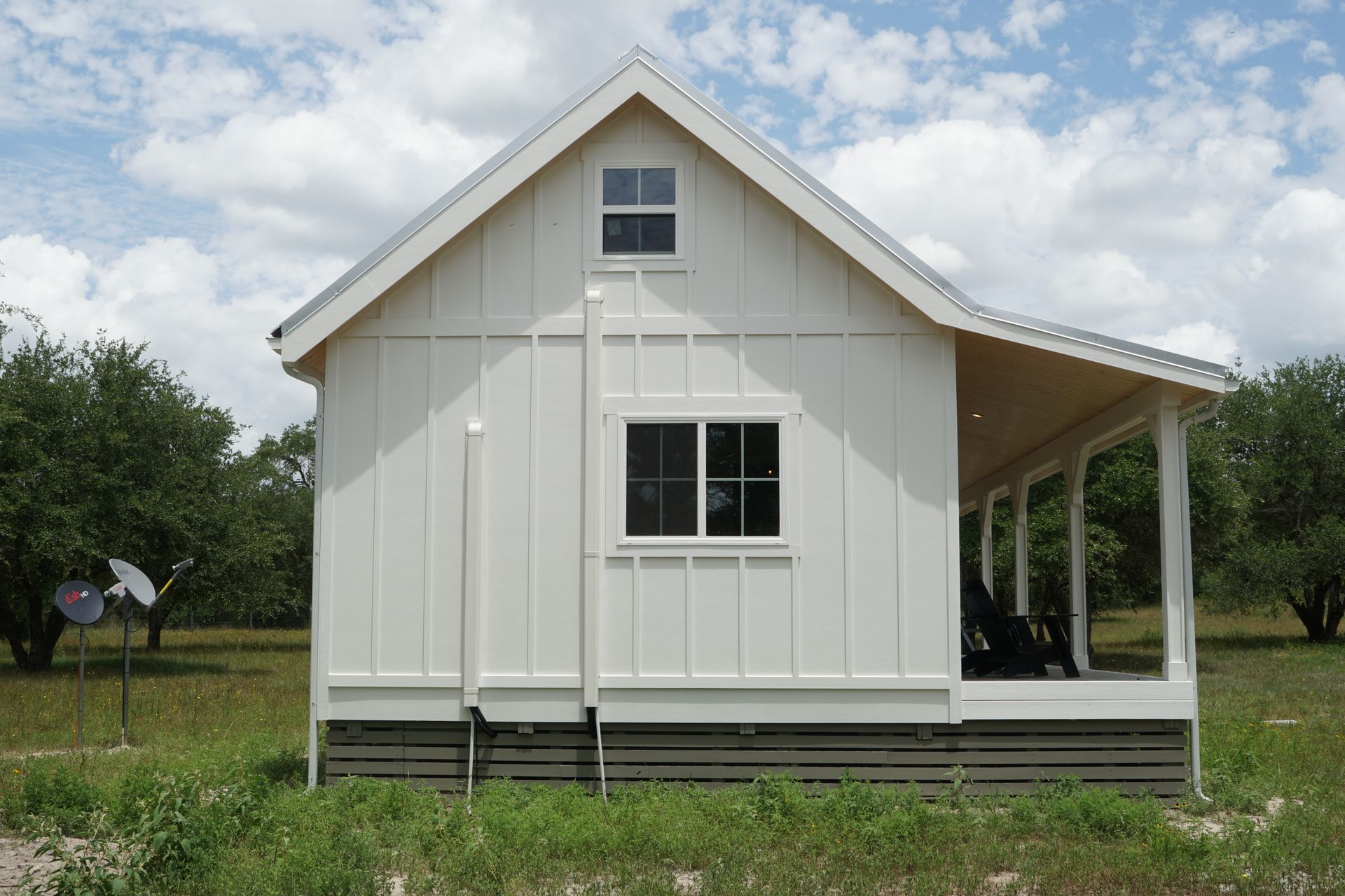 White cabin with porch, two windows, and vertical siding, set in a grassy field with trees under a cloudy sky.