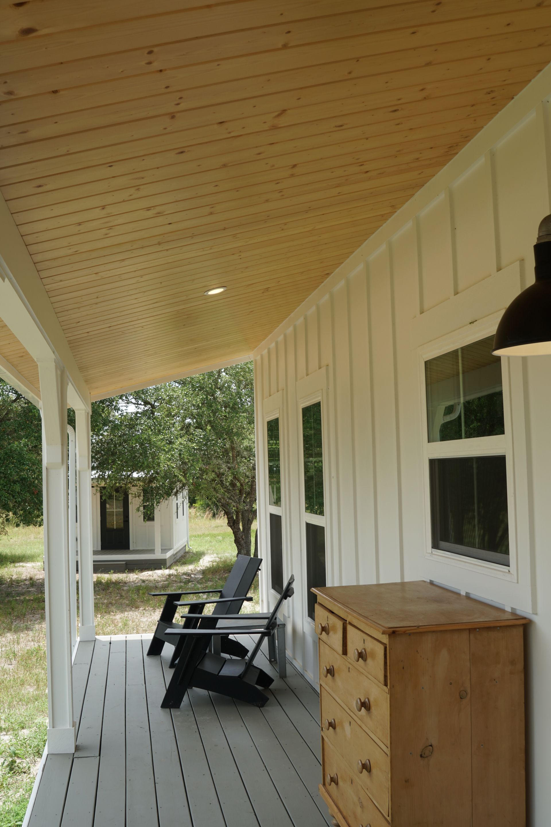 A white porch with gray flooring, wood ceiling, and a dresser. Black chairs sit in the distance.