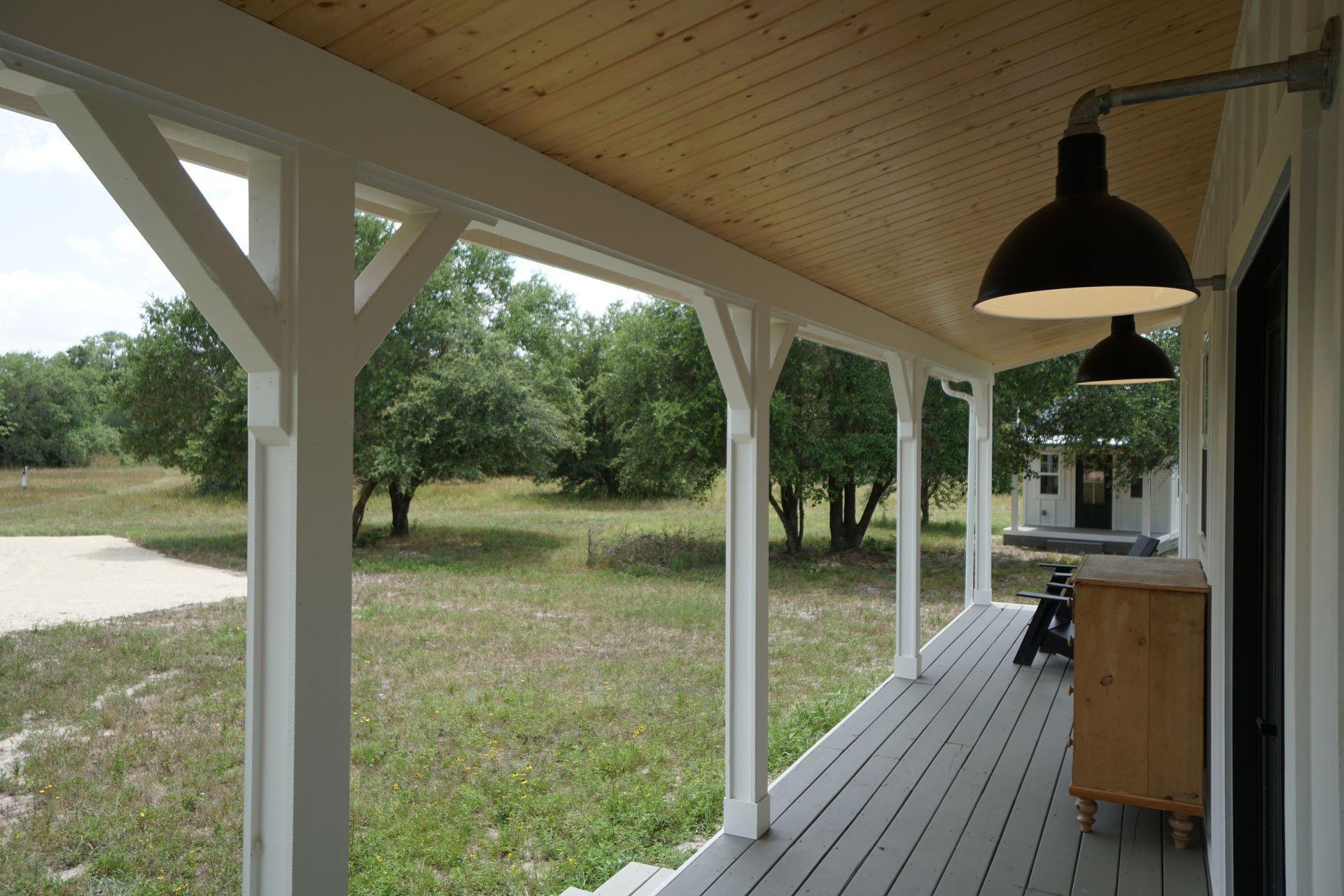 Covered porch with white columns, gray deck, and black overhead lights overlooking a grassy field with trees.