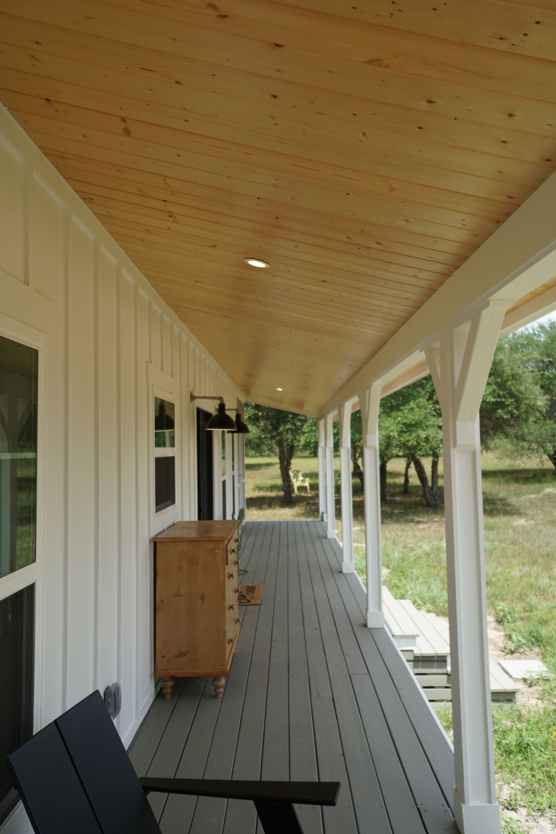 Covered porch with white siding, wooden ceiling, and gray floor. A wooden dresser stands near a black chair.