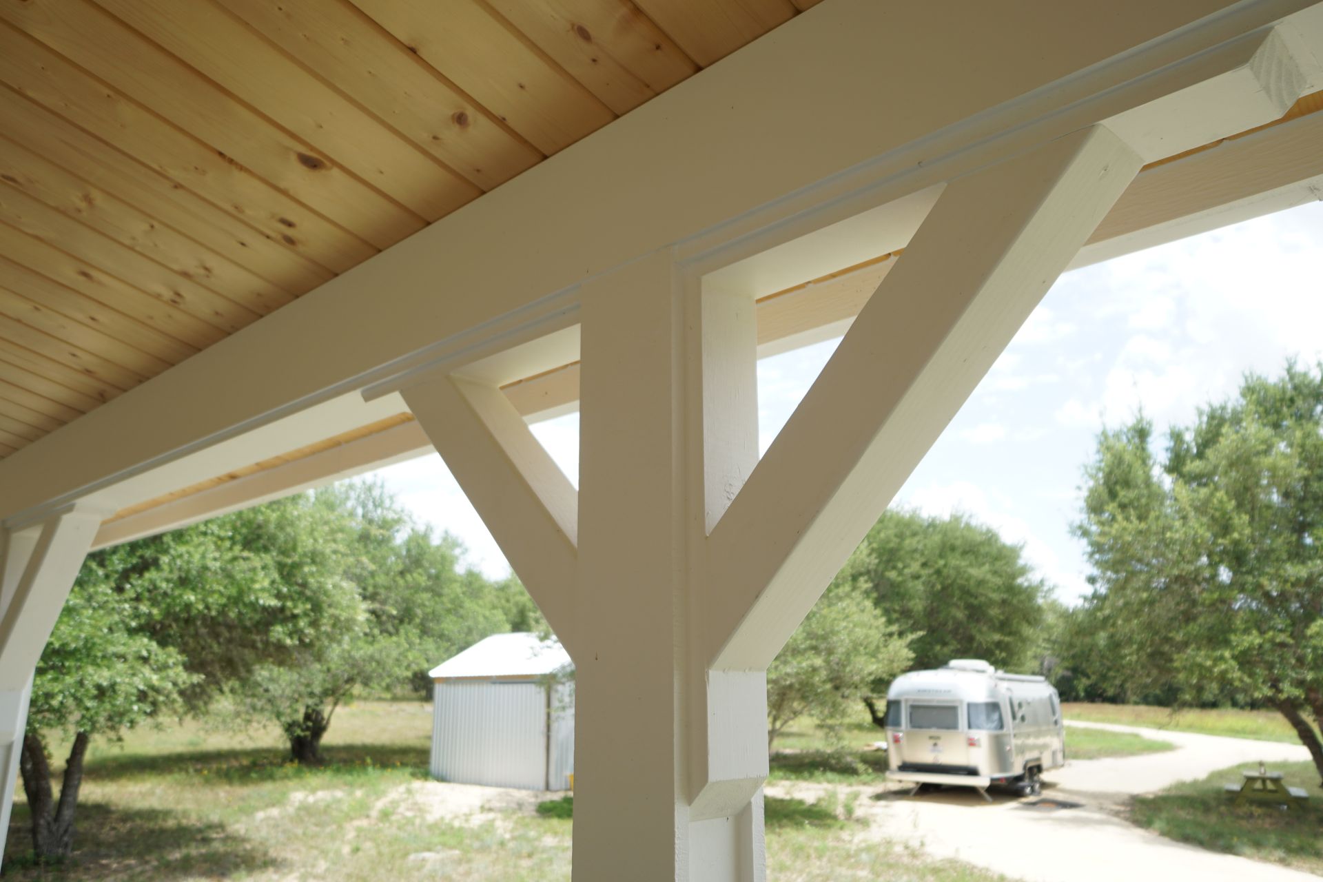 White porch with wooden ceiling, framing a view of a road, trees, and an Airstream trailer.